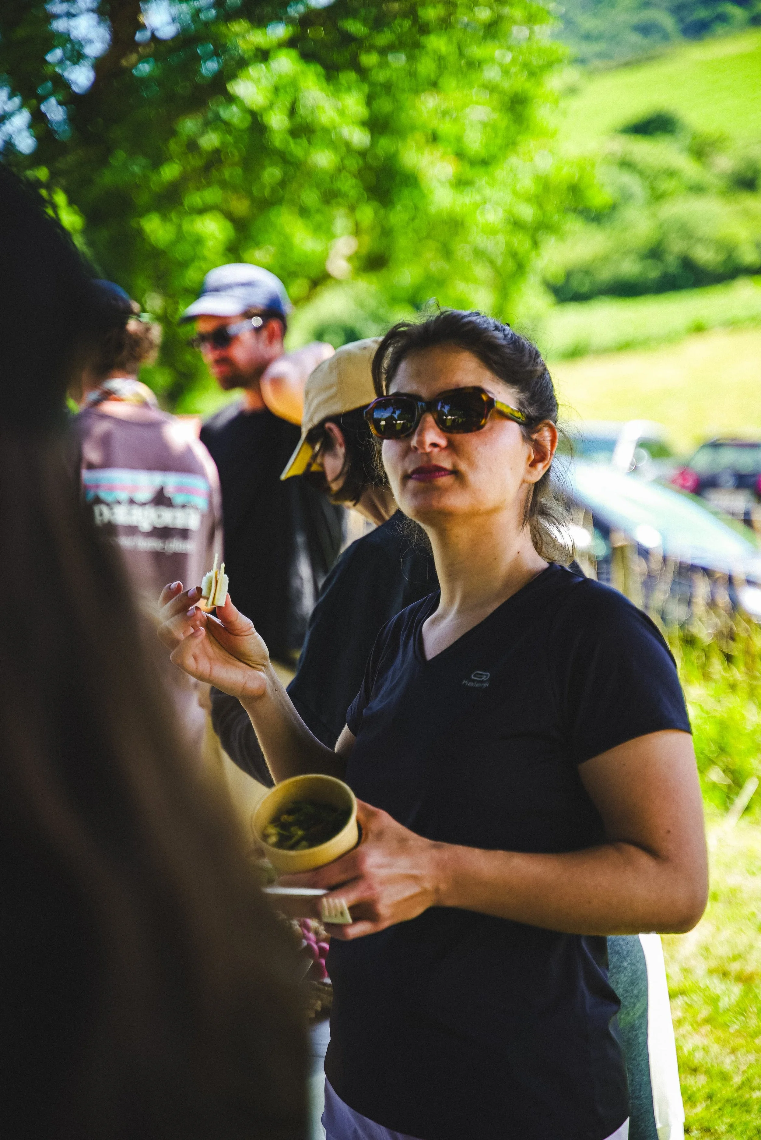 Une femme portant des lunettes de soleil noires, une chemise noire et tenant une tasse jaune en regardant vers la caméra, lors d'un événement en plein air avec des personnes en arrière-plan et un paysage verdoyant.