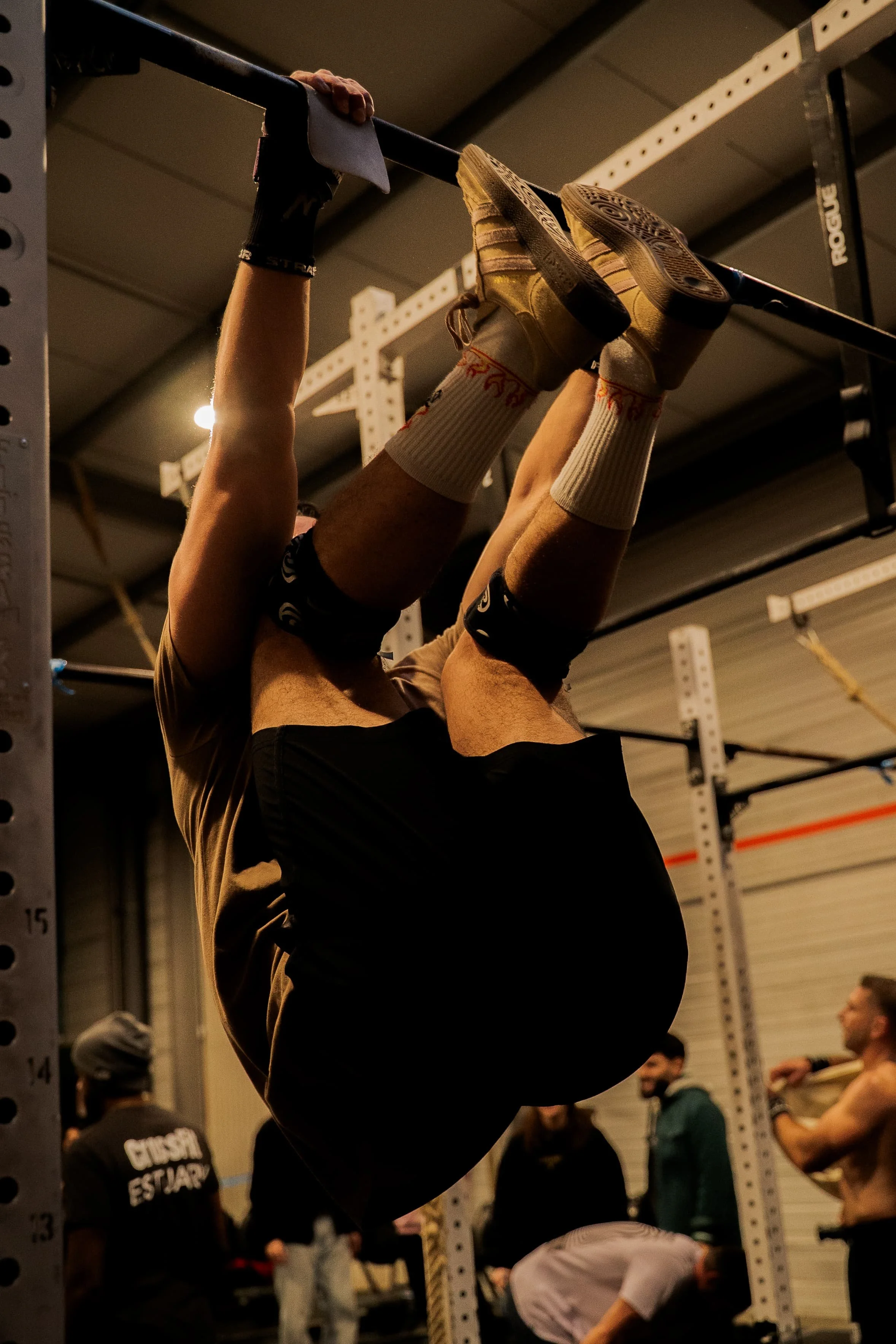 Une personne est suspendue à une barre de traction, en train de faire un exercice de musculation, dans une salle de sport. Plusieurs autres personnes sont visibles en arrière-plan, certaines assises ou debout.