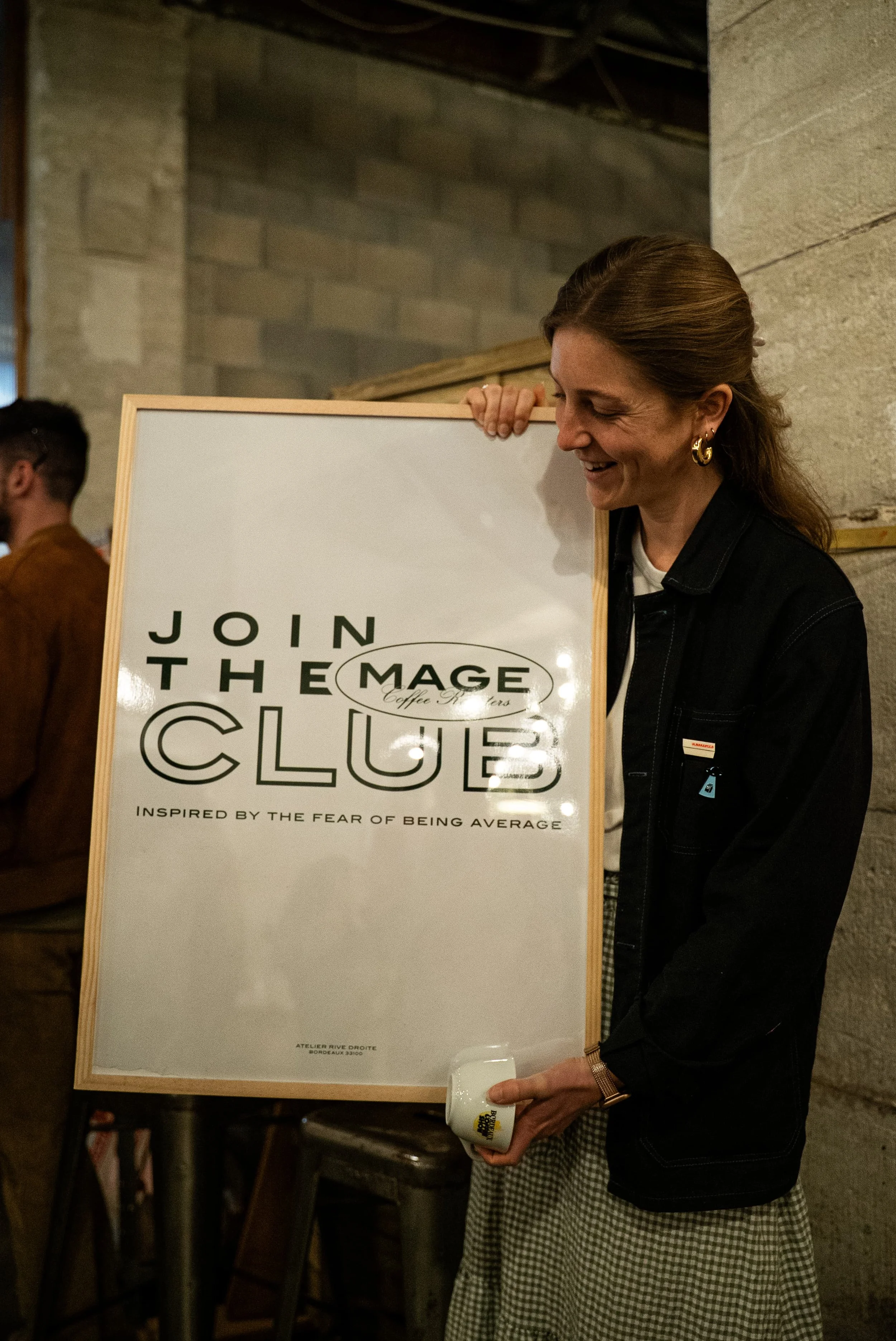 Une femme souriante qui tient une affiche avec le texte 'Rejoignez le club'. Elle porte des boucles d'oreilles en or et une montre. L'environnement semble être un lieu intérieur, probablement un atelier ou une galerie.
