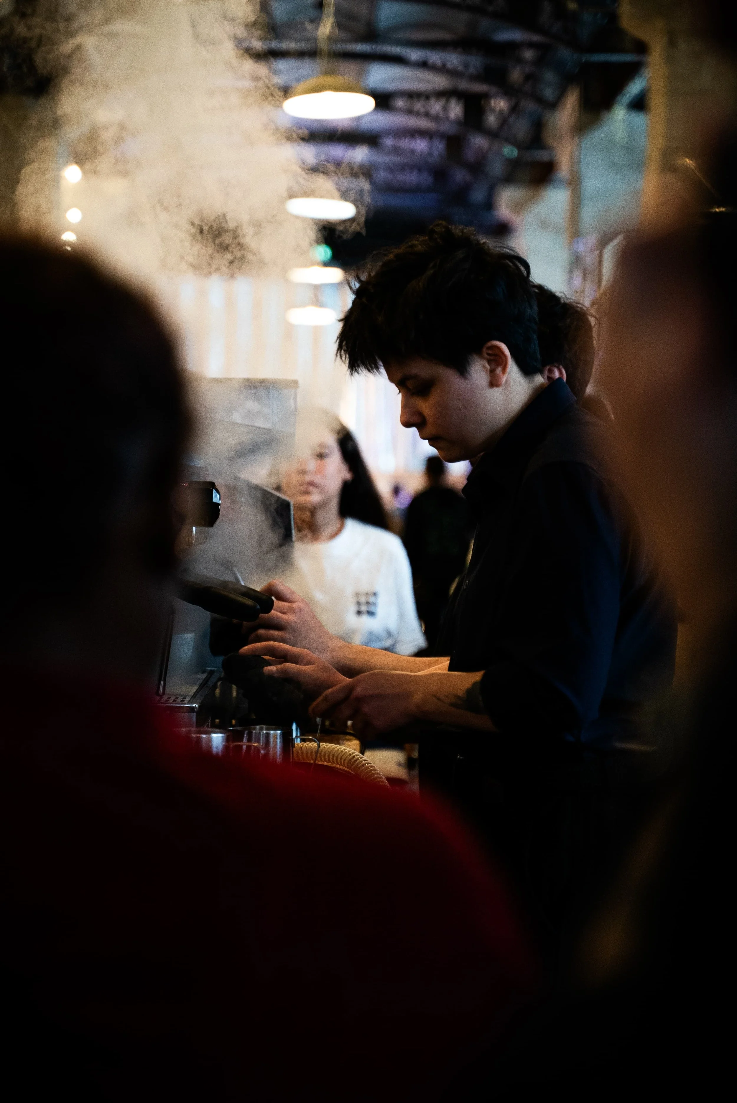 Une personne avec des cheveux noirs courts, portant une chemise noire, se trouve derrière un comptoir, probablement dans un café ou restaurant, utilisant un terminal ou une caisse. Des clients et autres personnes sont visibles en arrière-plan dans un