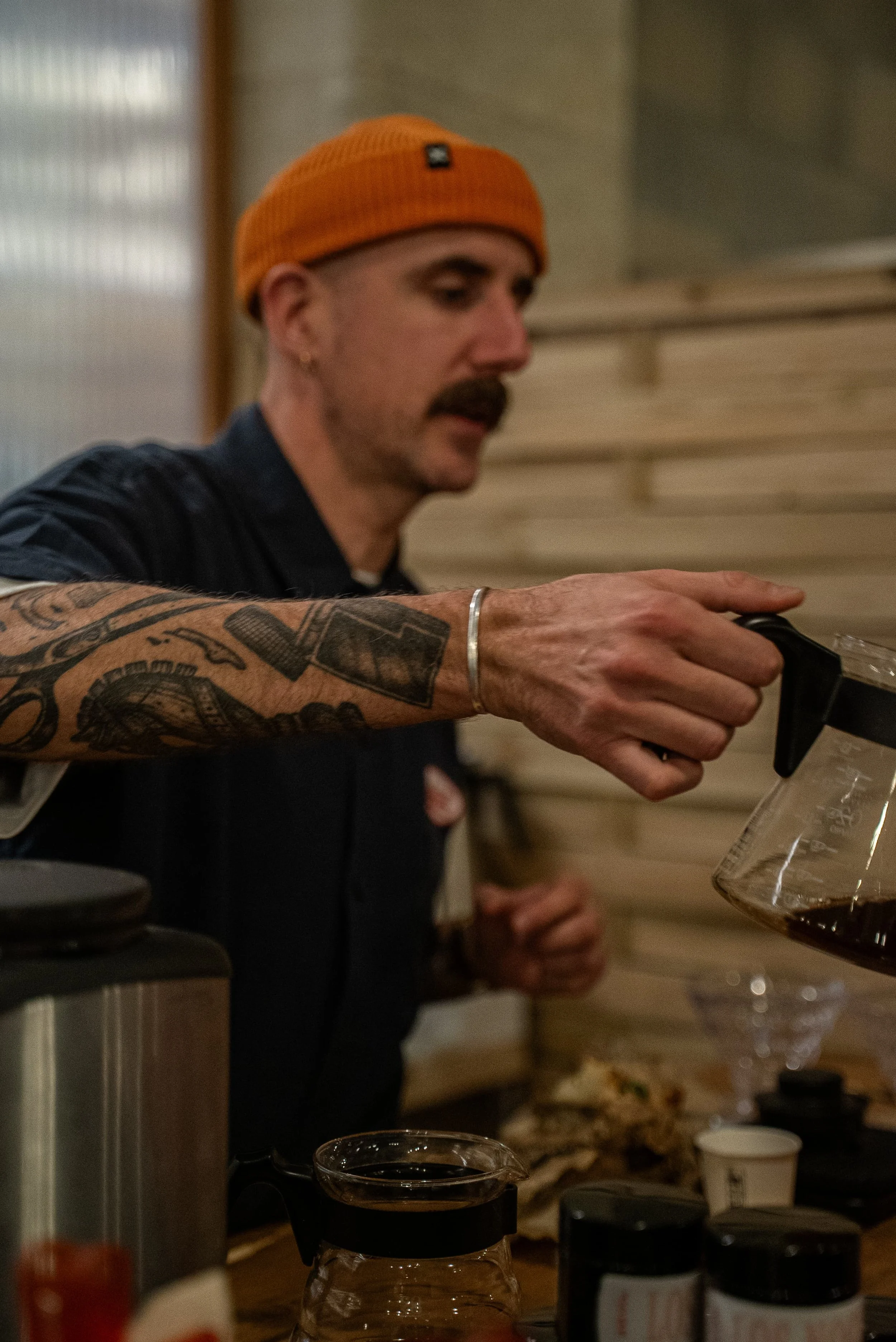 Un homme portant un bonnet orange verse du café dans une cafetière en verre. Autres éléments comme un touret, plusieurs bocaux et de la nourriture sont visibles en arrière-plan.