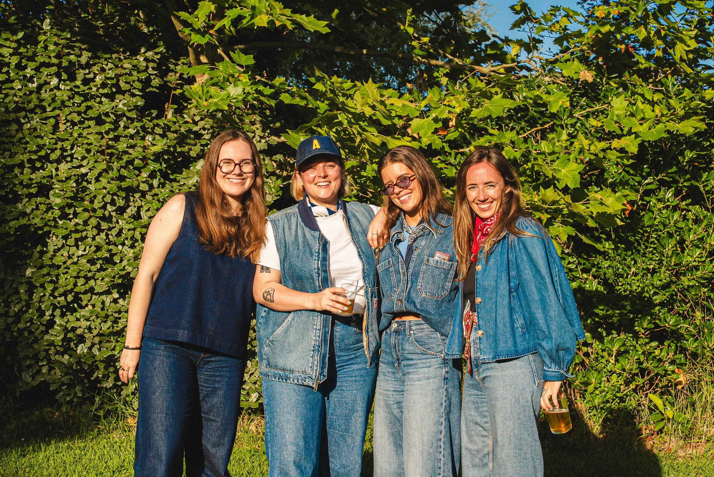 Groupe de quatre femmes souriantes, habillées en denim, posant devant un mur de feuillage vert avec un ciel bleu en arrière-plan, lors d'une journée ensoleillée, tenant un verre de bière.
