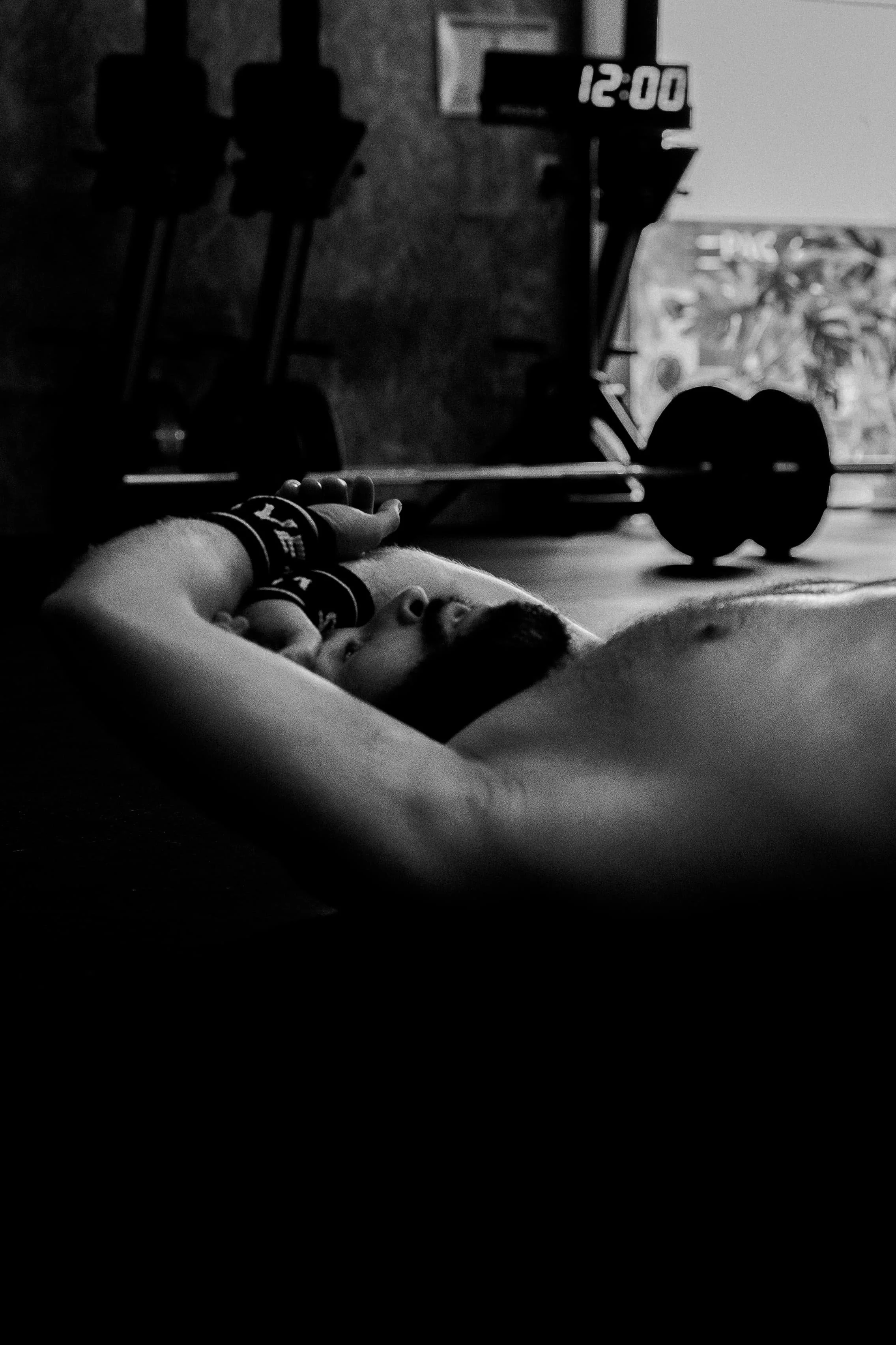 Un bras musclé allongé avec un bandeau noir, à côté d'une paire de haltères, dans une salle de sport avec un affichage numérique de l'heure à 12h00 en arrière-plan.