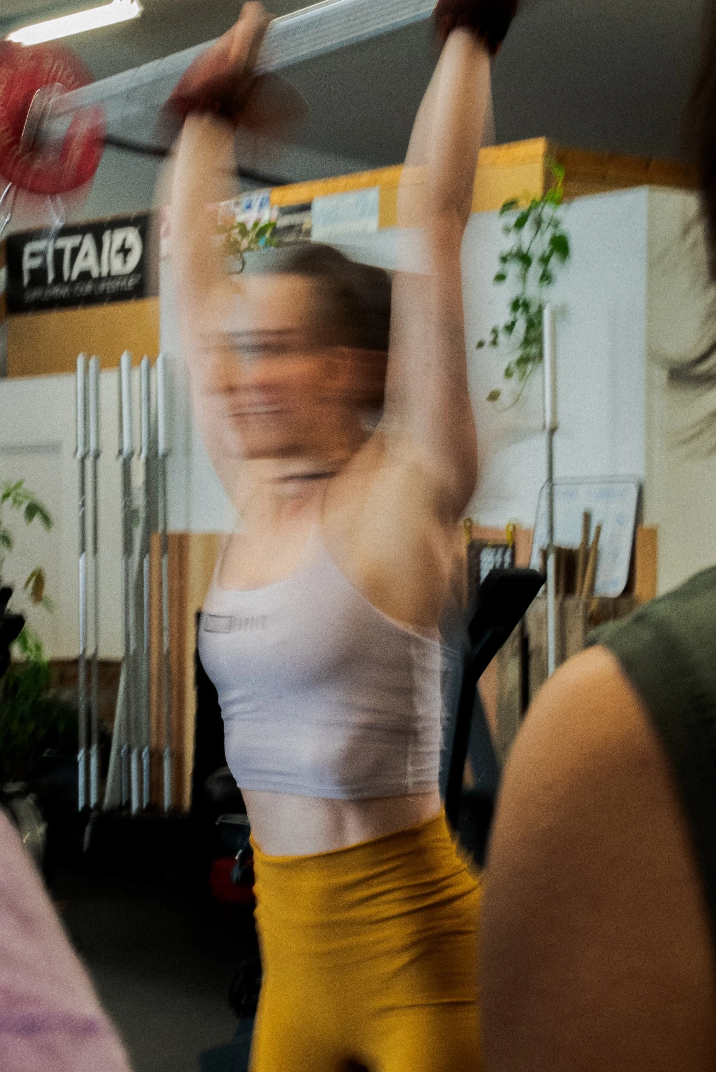 Une femme en tenue de sport jaune et blanc en train de faire des exercices ou de s'étirer dans une salle de sport ou de fitness. La photo est prise dans un environnement intérieur, avec des plantes et des équipements de sport en arrière-plan.