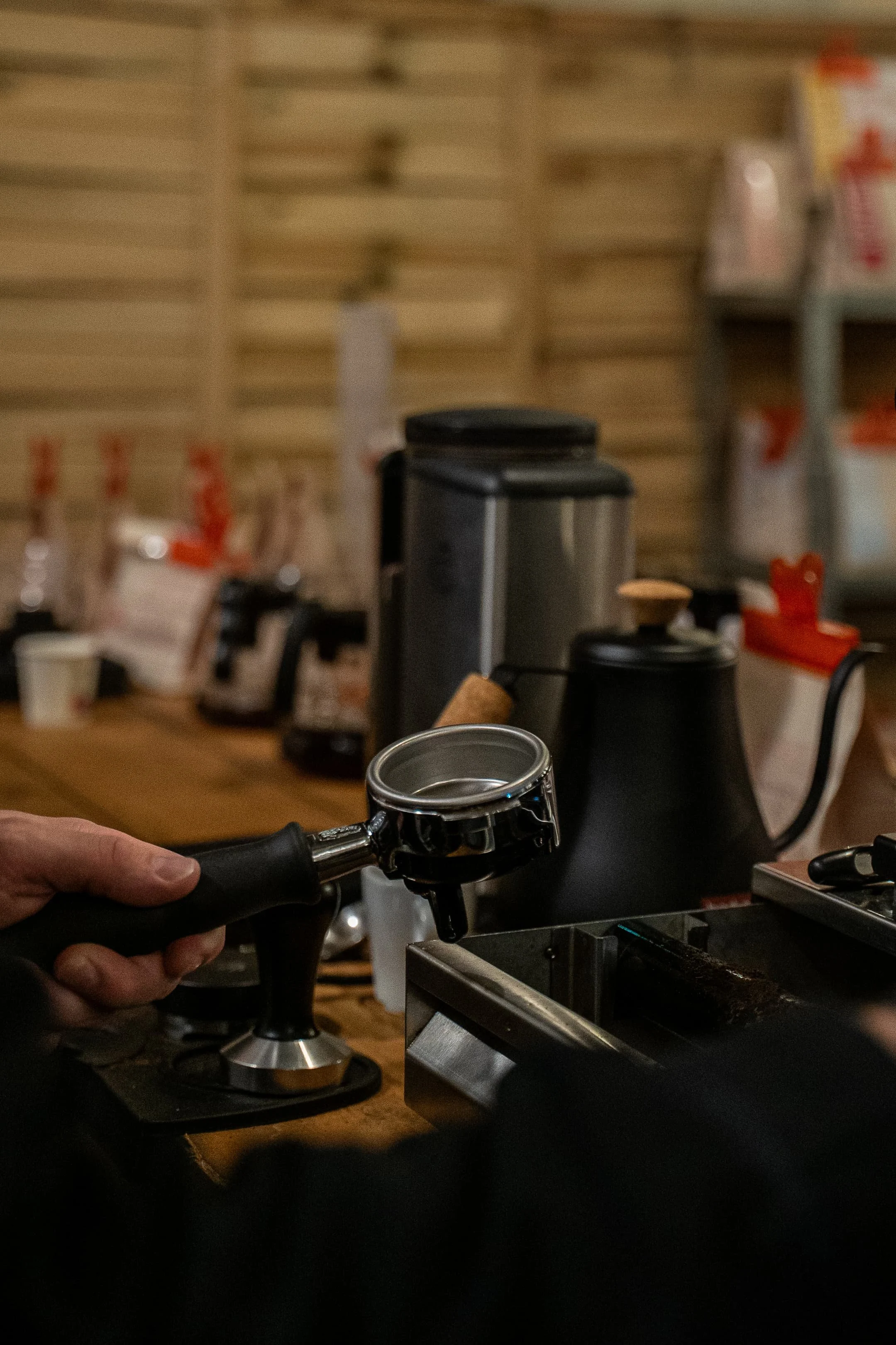Une personne tient un manche de machine à espresso noire, prête à l'utiliser sur une machine à café dans une cuisine avec étagères en bois et divers paquets de café ou d'autres produits en arrière-plan.