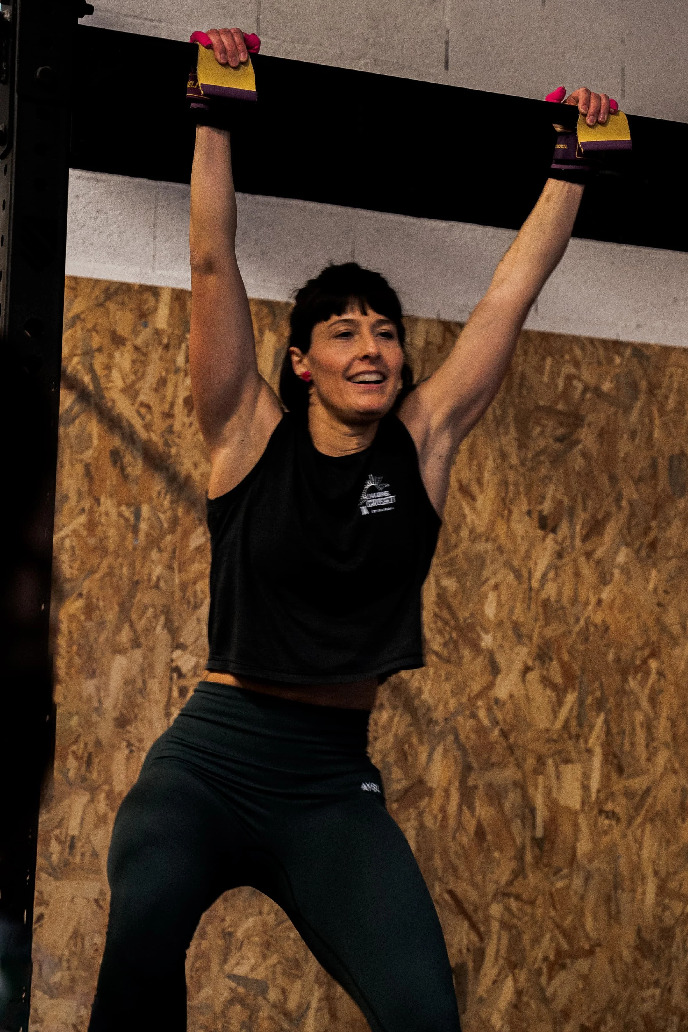 Une femme fait de l'exercice, elle se tient debout, les bras levés tenant une barre de traction, dans une salle d'entraînement avec un mur en panneau de bois.