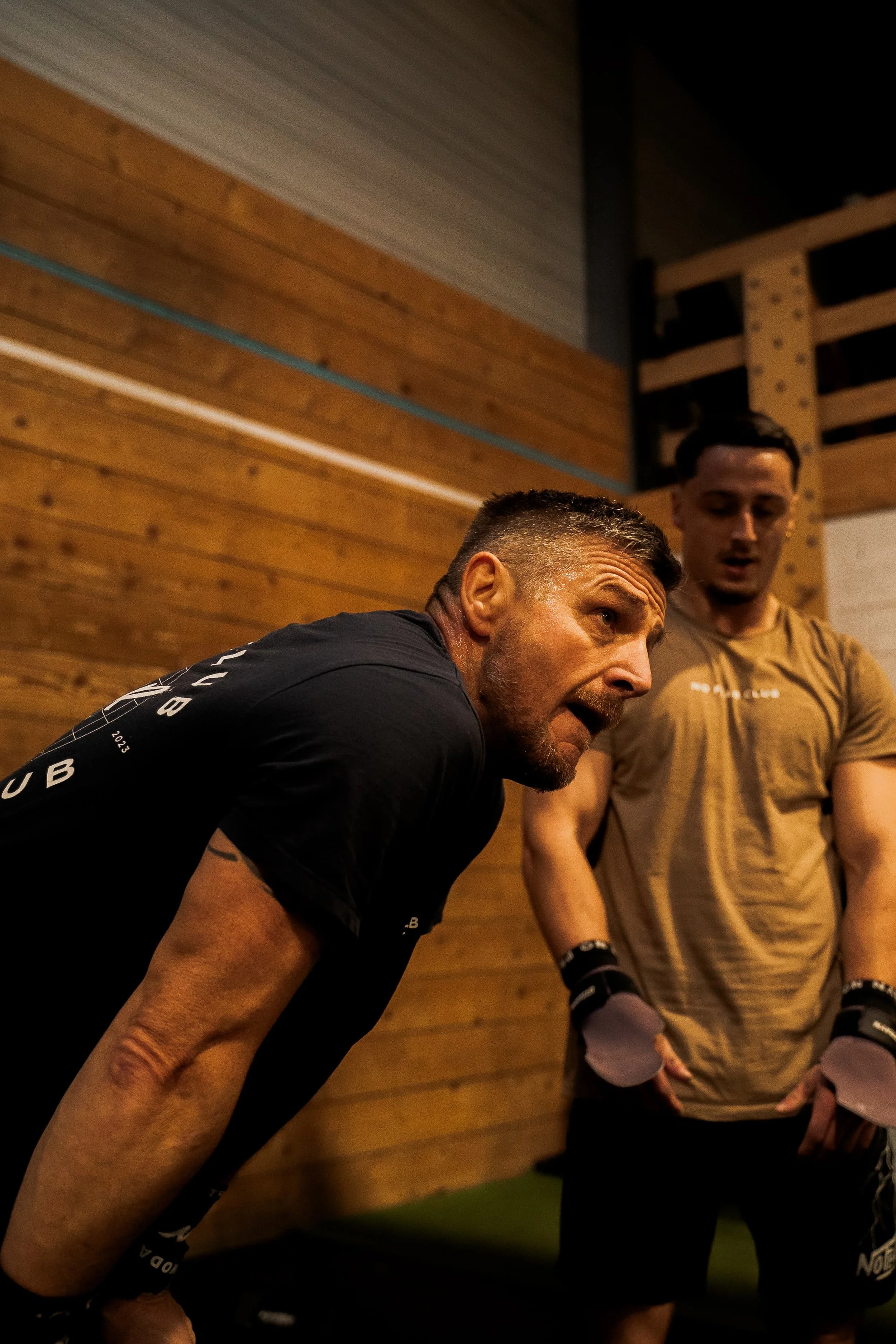 Deux hommes en vêtements de sport, concentrés, semblent en pleine discussion ou entraînement dans une salle à bois avec un mur en panneaux et un plafond en métal.