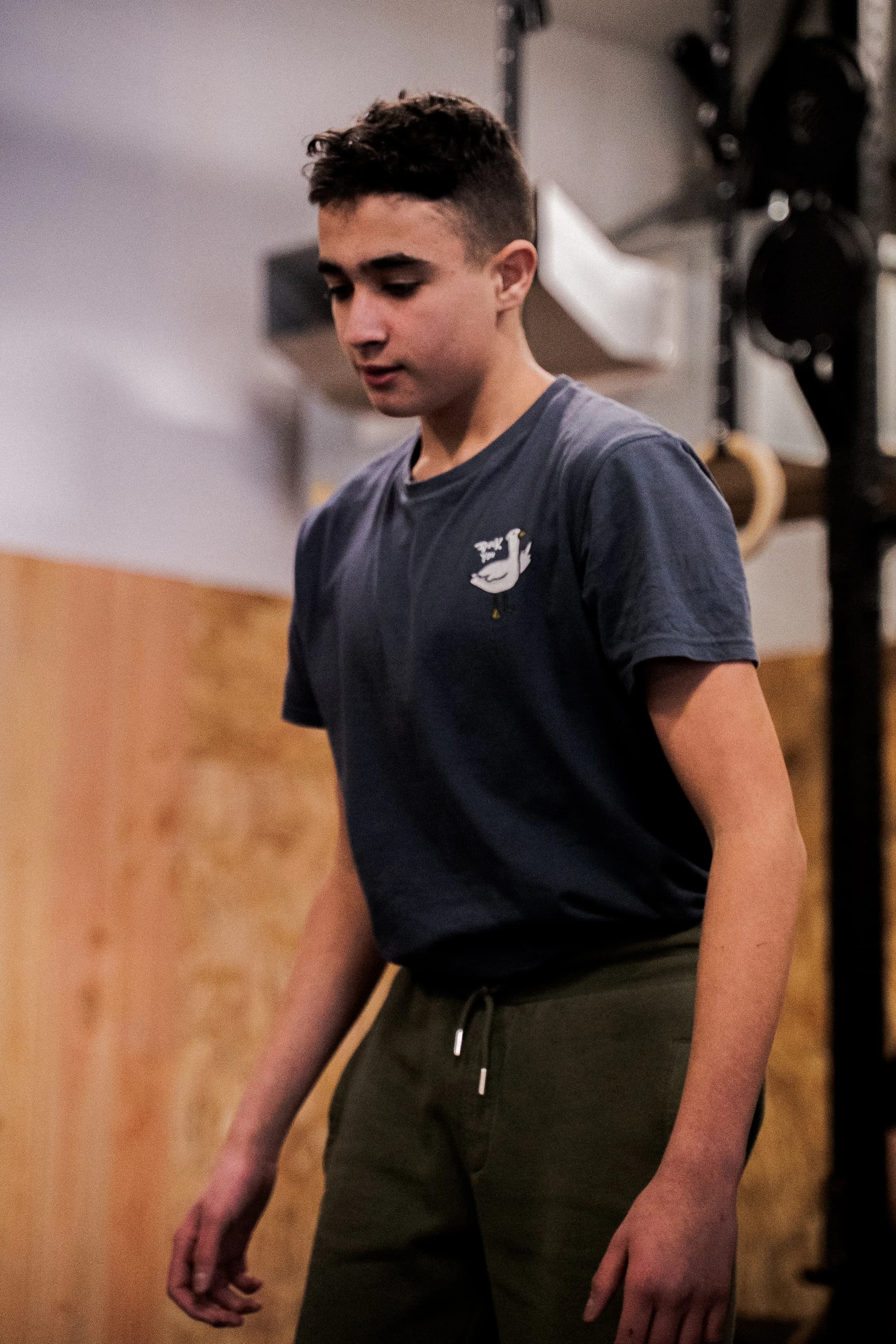 Jeune homme portant un t-shirt gris avec un dessin de cygne, en salle de musculation, posture détendue.