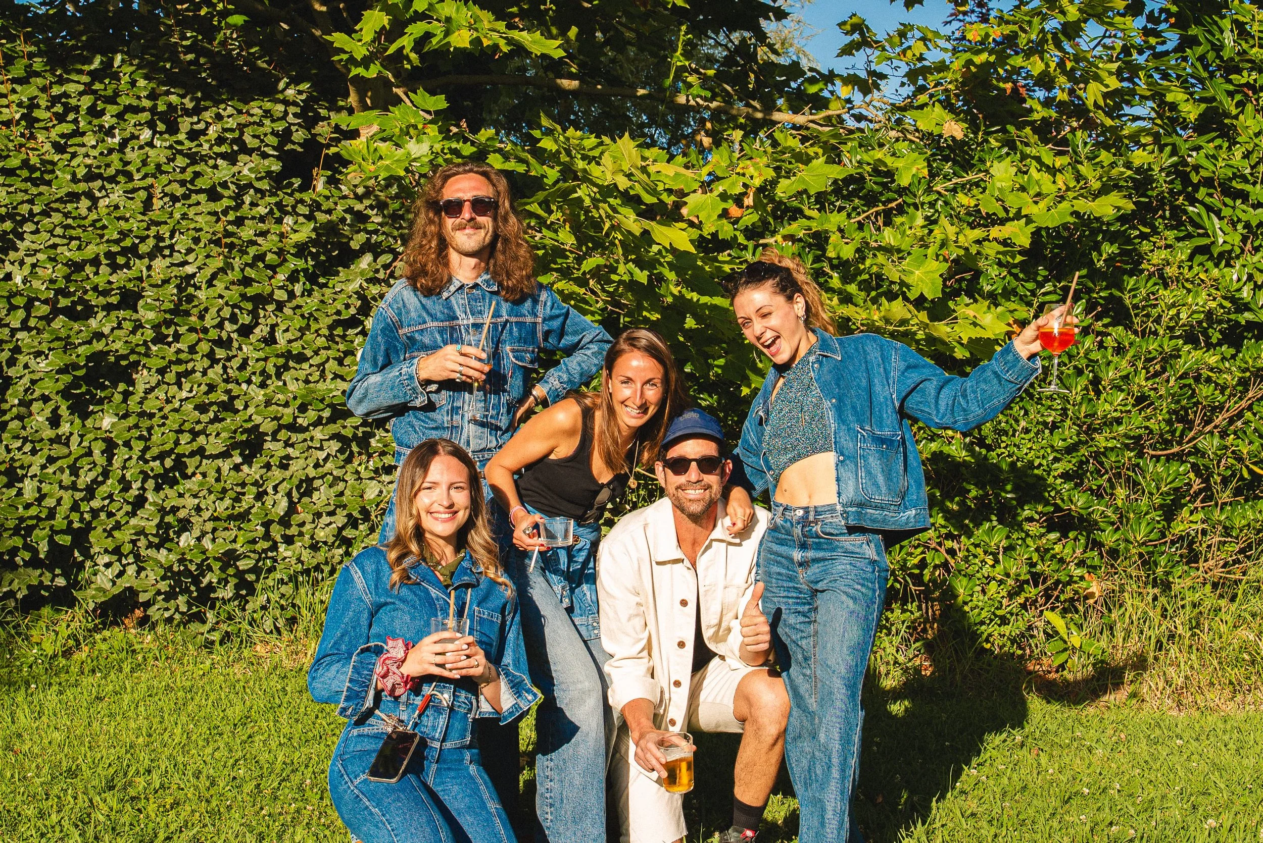 Groupe de personnes posant pour une photo en plein air, entouré de verdure, certains tenant des verres de boissons alcoolisées.