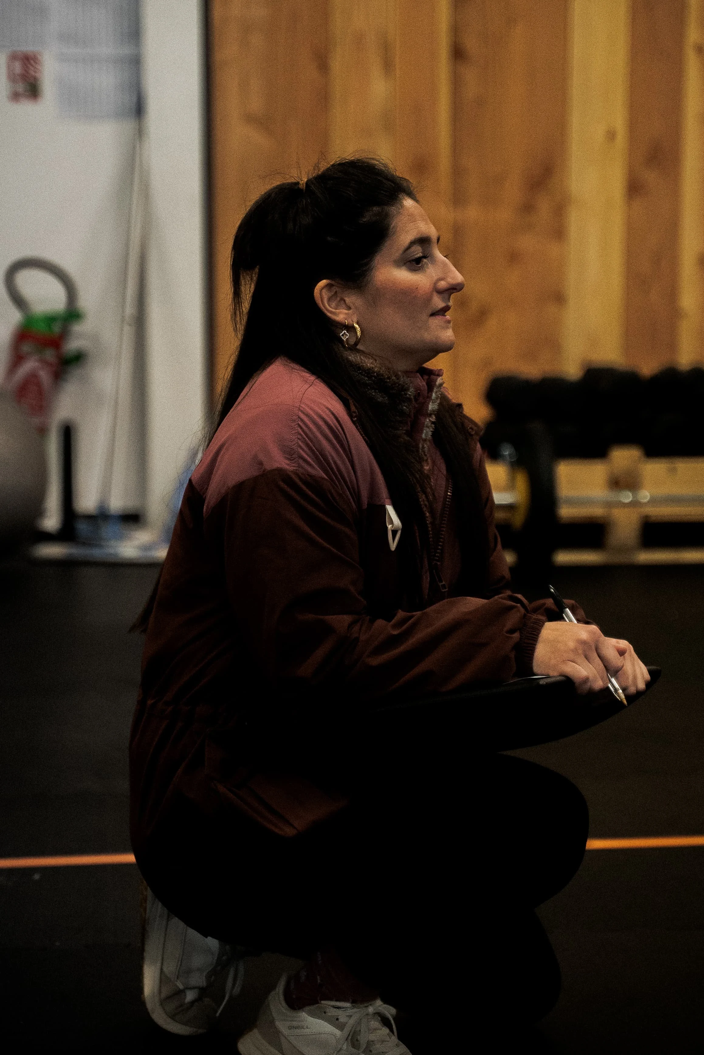 Une femme assise dans une salle en bois, portant une veste marron et des sneakers blanches, en train d'écrire avec un stylo sur une surface noire.