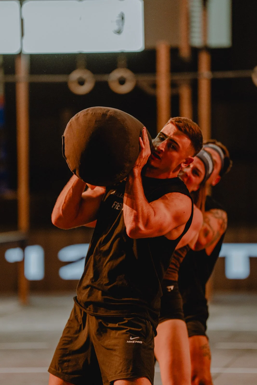 Homme soulevant une medecine ball lors d'une séance CrossFit avec deux athlètes en arrière-plan à Bordeaux — photographe CrossFit Voar by Tim
