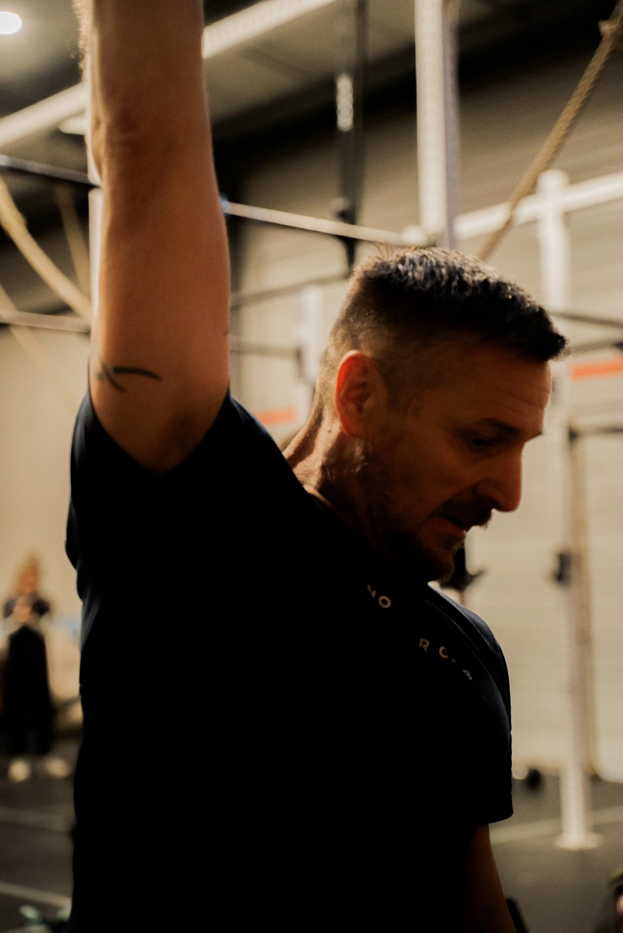 Un homme souriant dans une salle de sport ou une salle de fitness, portant un t-shirt noir, en train de faire de l'exercice ou de s'étirer.