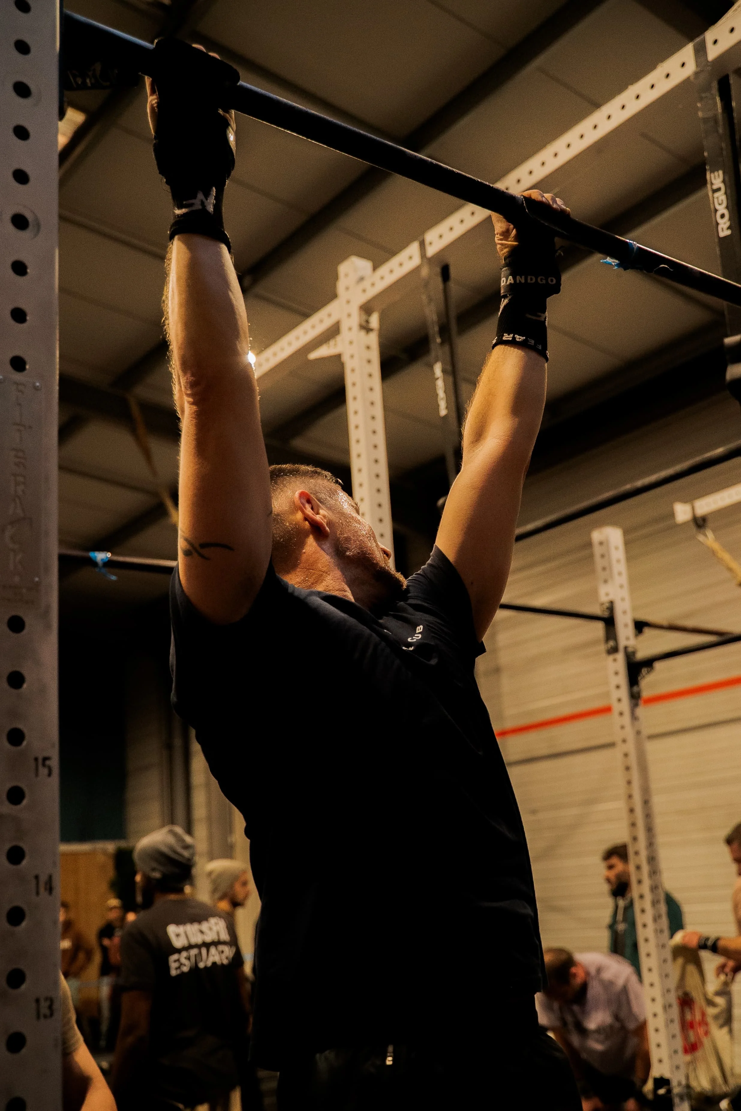 Une personne effectuant un exercice de traction à la barre dans une salle de sport, plusieurs autres personnes en arrière-plan.