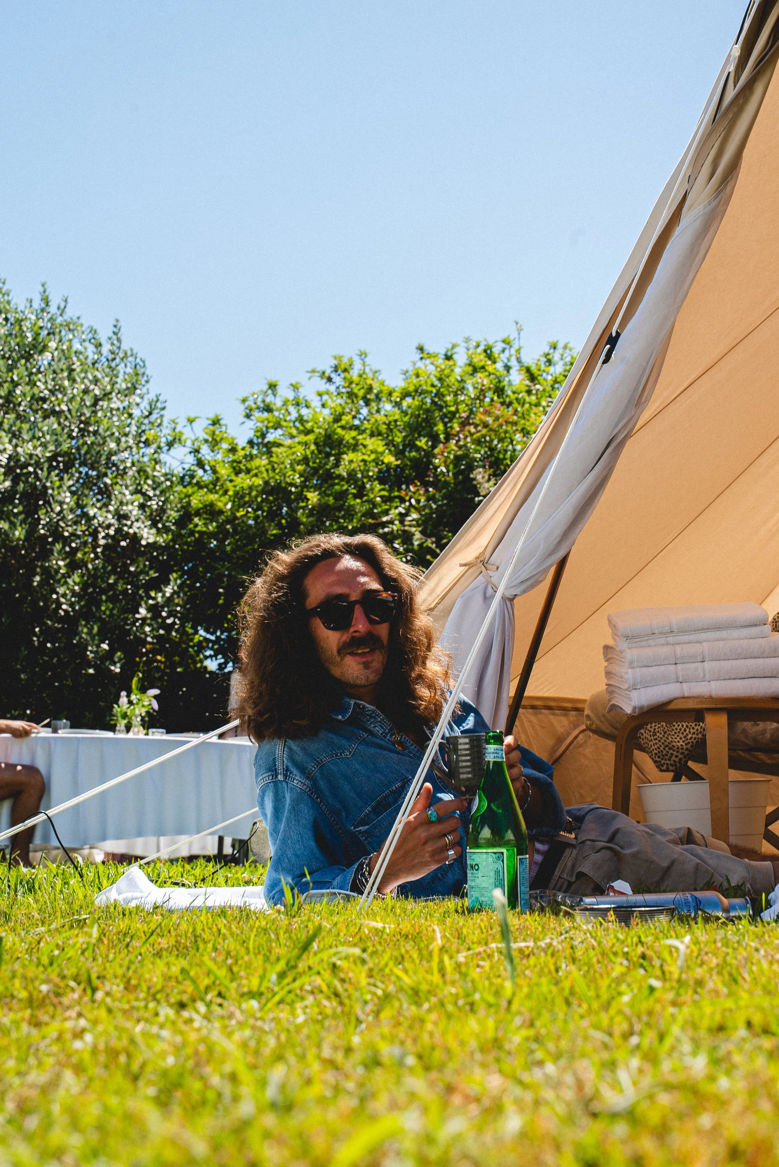 Homme à lunettes avec cheveux longs et barbe, assis sur l'herbe près d'une tente beige, tenant une bouteille verte, dans un jardin ensoleillé avec des arbres en arrière-plan.