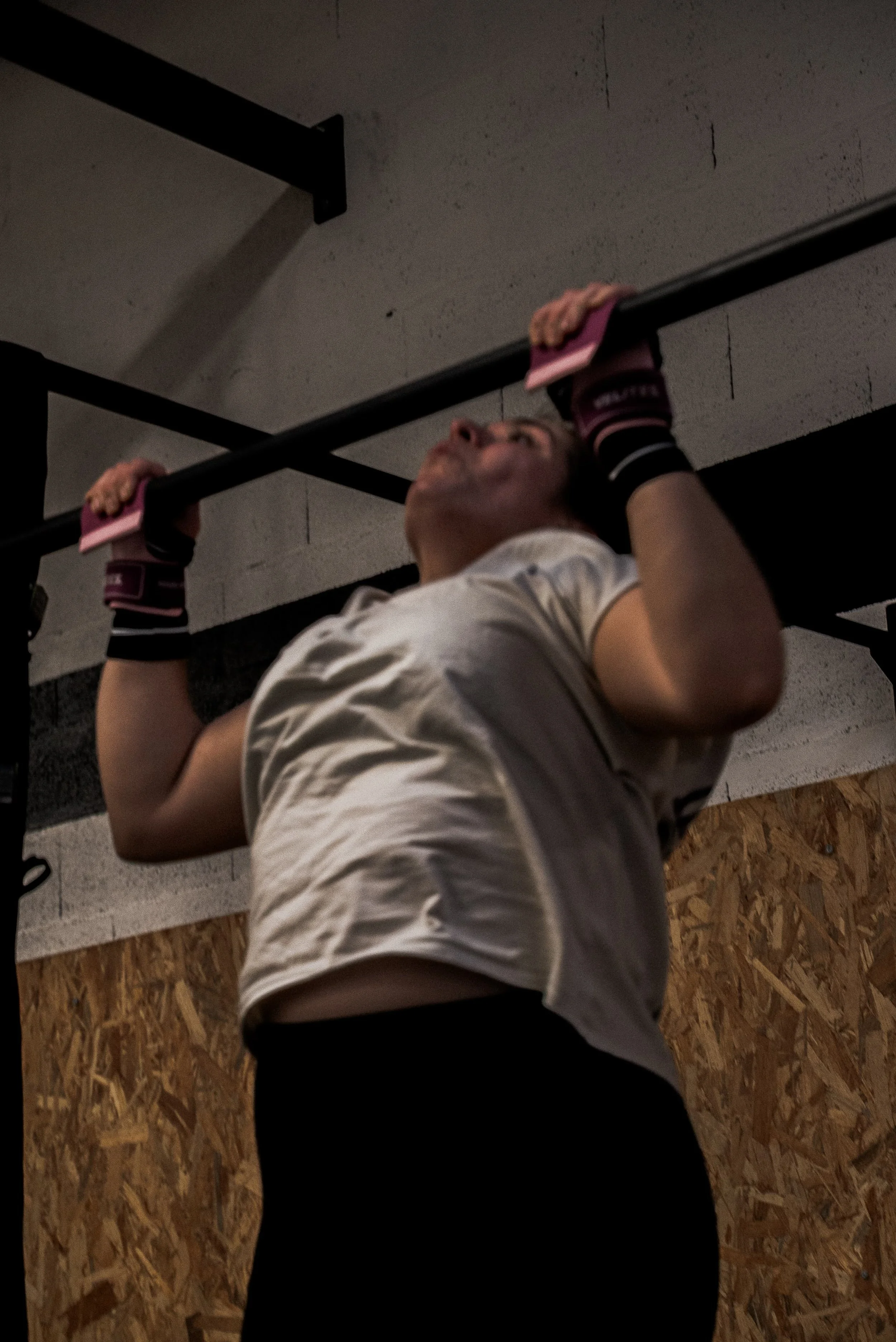 Une personne effectue une traction à la barre dans une salle de sport. La personne porte des gants de musculation et un t-shirt blanc, avec un mur en bois en arrière-plan.