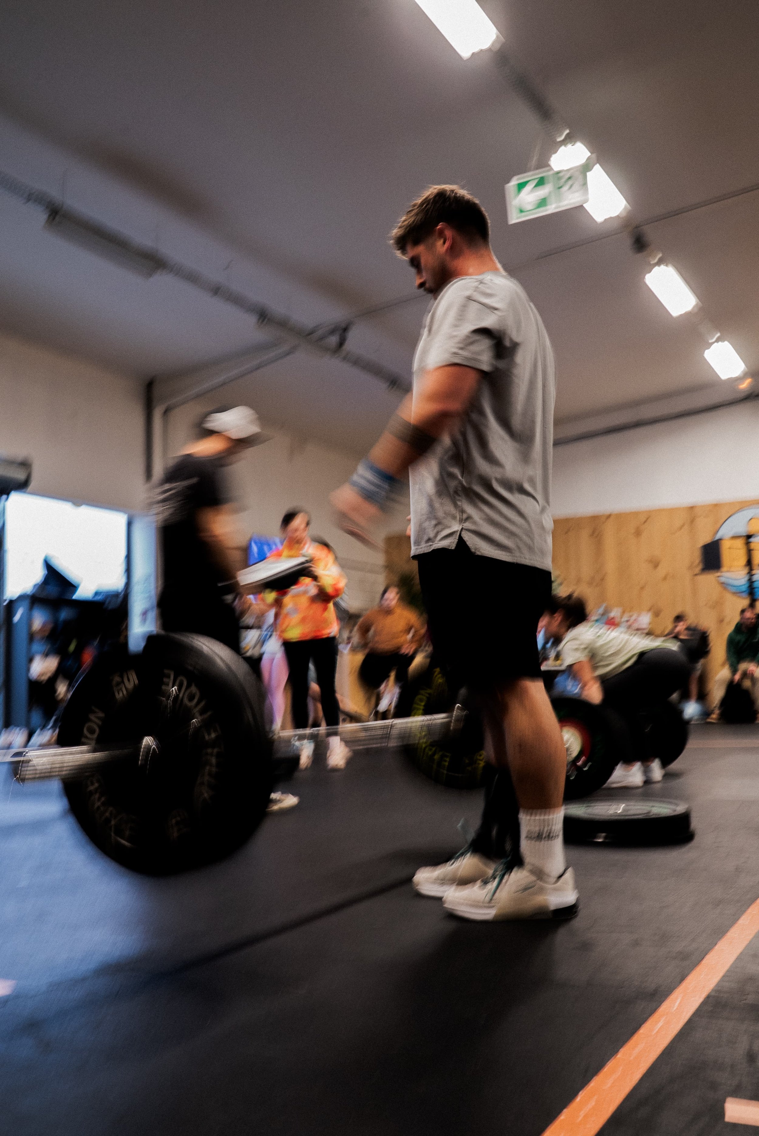 Jeune homme en vêtements de sport, portant des chaussettes de sport, effectuant un soulevé de terre avec une barre de poids dans une salle de sport. Autres personnes en arrière-plan, certains préparant des exercices ou parlant, dans une ambiance dyna
