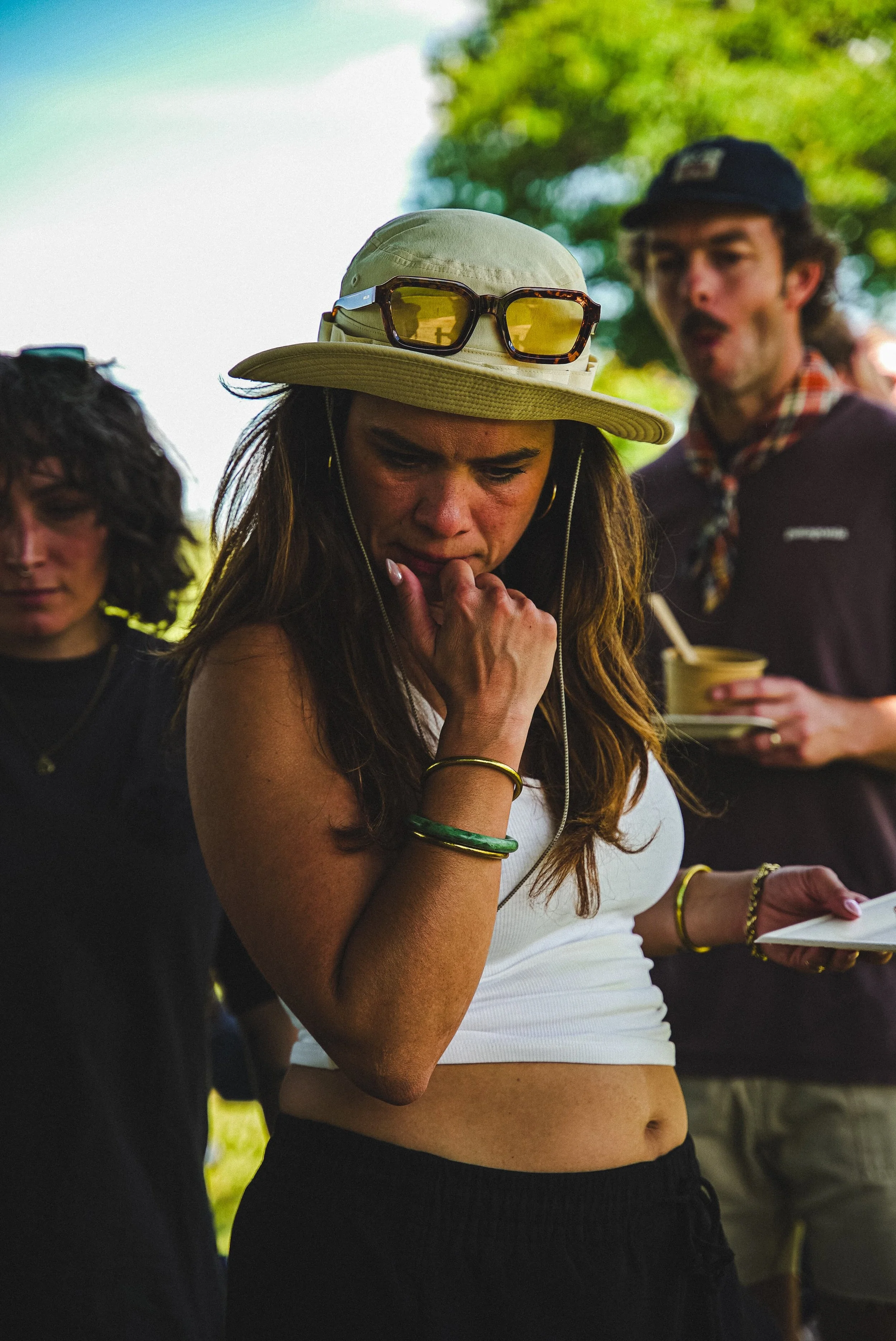 Une femme portant un chapeau beige avec des lunettes de soleil, vêtue d'un haut blanc et de pantalons noirs, avec plusieurs bracelets en or et en plastique, semble être en train de réfléchir ou d'observer quelque chose lors d'un événement en plein ai
