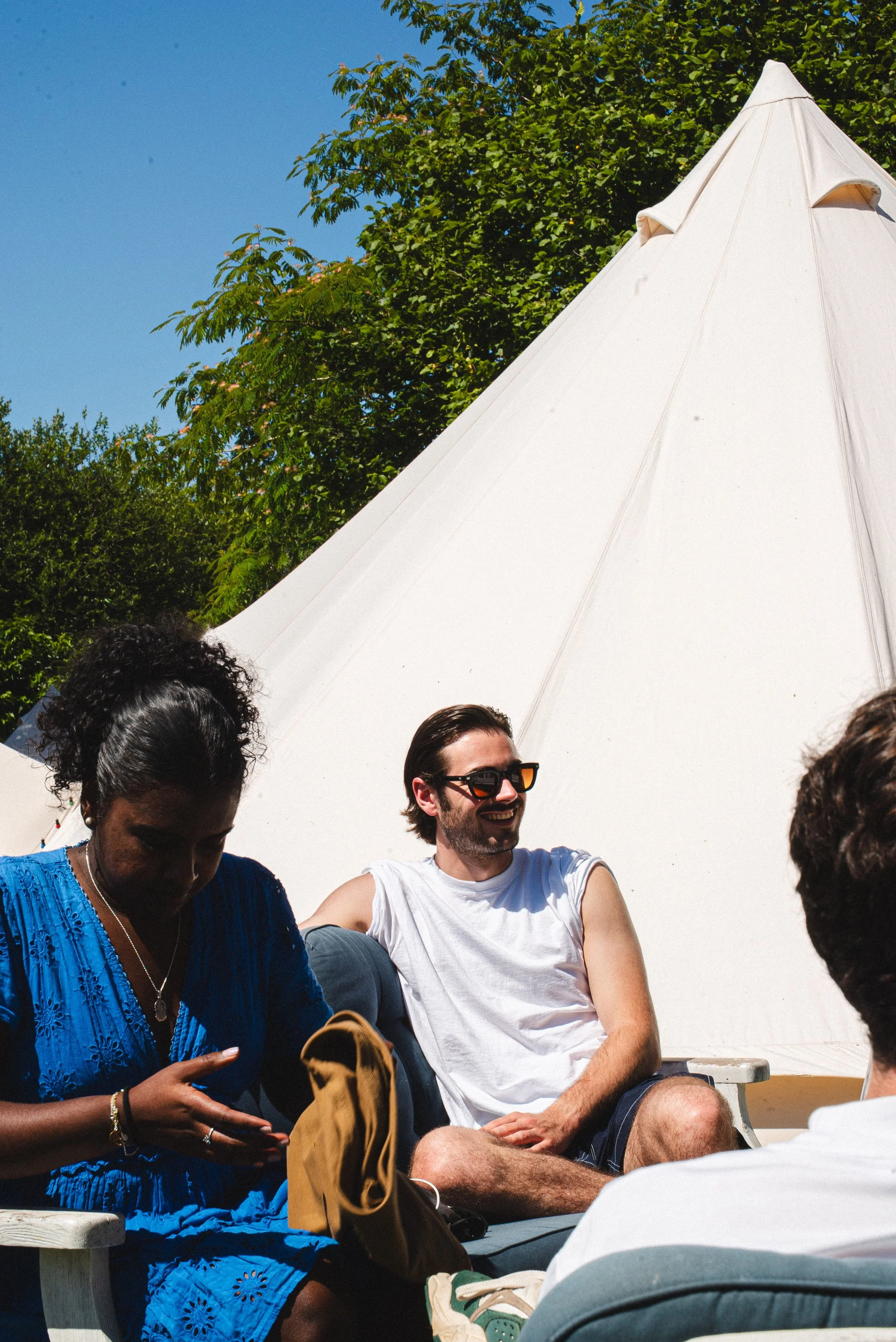 Groupe de personnes assises sous un parasol blanc lors d'une journée ensoleillée, avec des arbres verts en arrière-plan.
