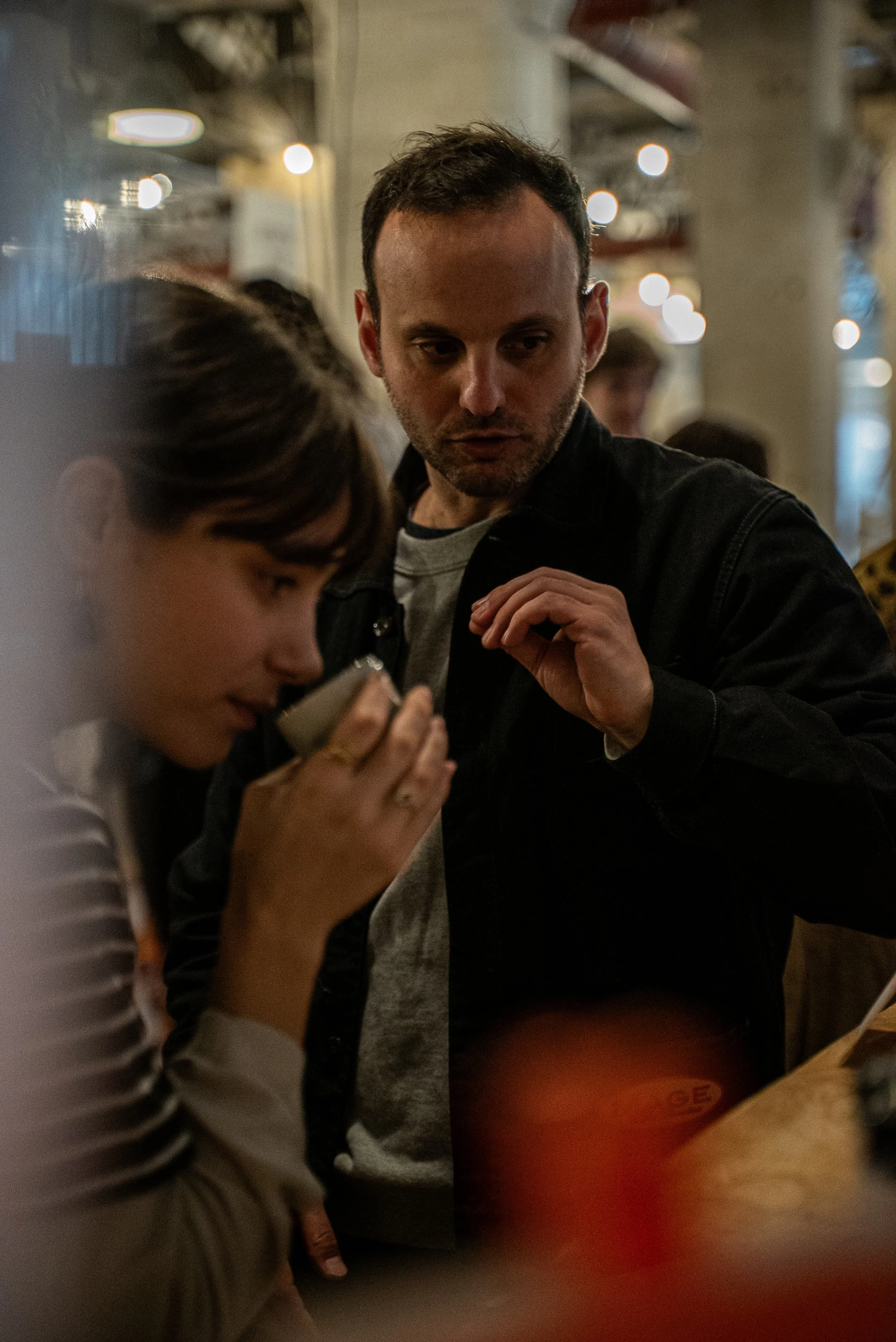 Deux personnes discutent dans un endroit intérieur avec une ambiance chaleureuse et des lumières tamisées. La personne à droite a une expression sérieuse et regarde intensément, tandis que l'autre personne, à gauche, semble concentrée sur quelque cho