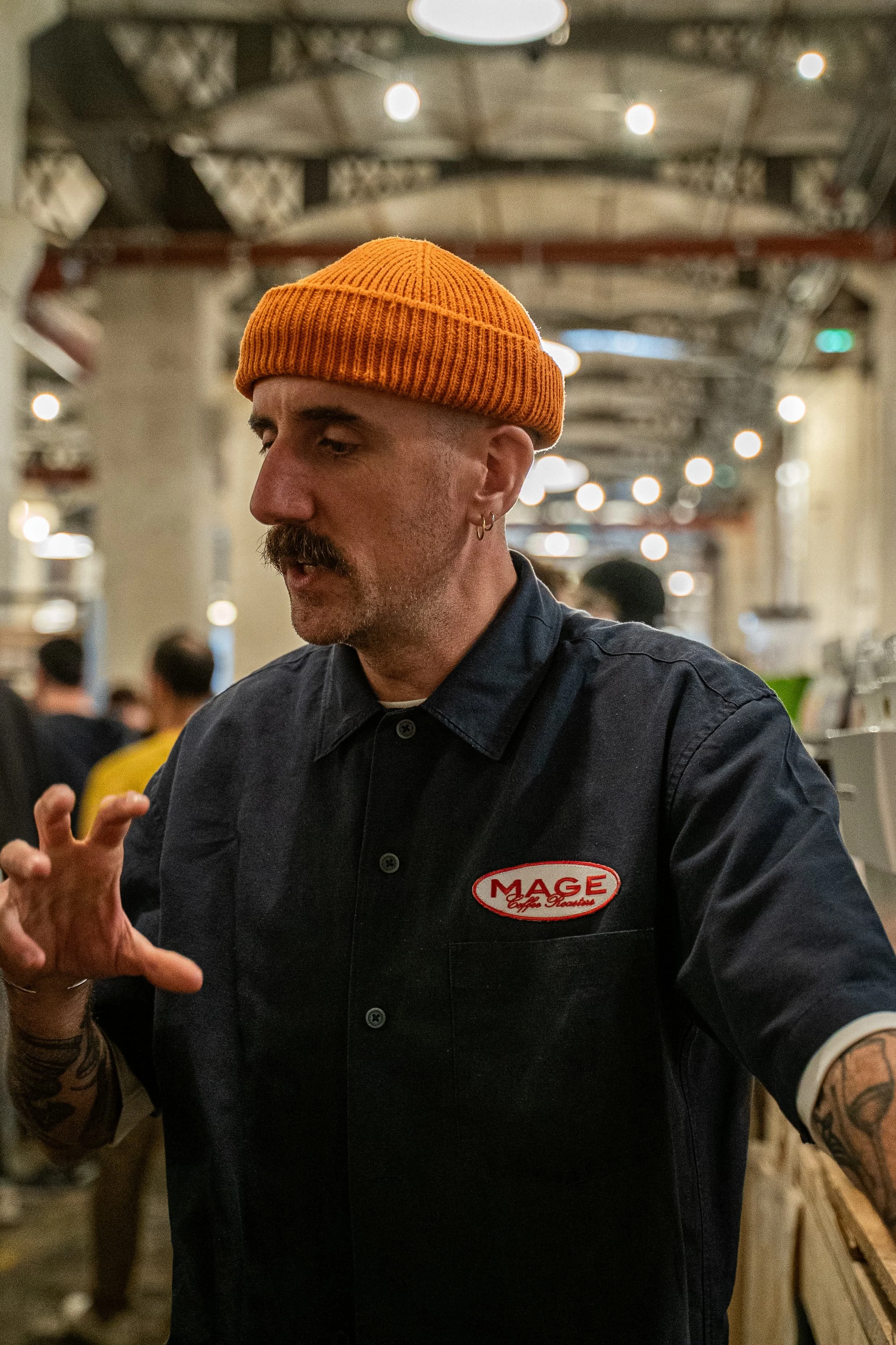 Homme portant un bonnet orange et un uniforme noir avec un logo « MAGE », discutant dans une ambiance de restaurant ou de café, avec des lumières suspendues au plafond et d'autres personnes en arrière-plan.