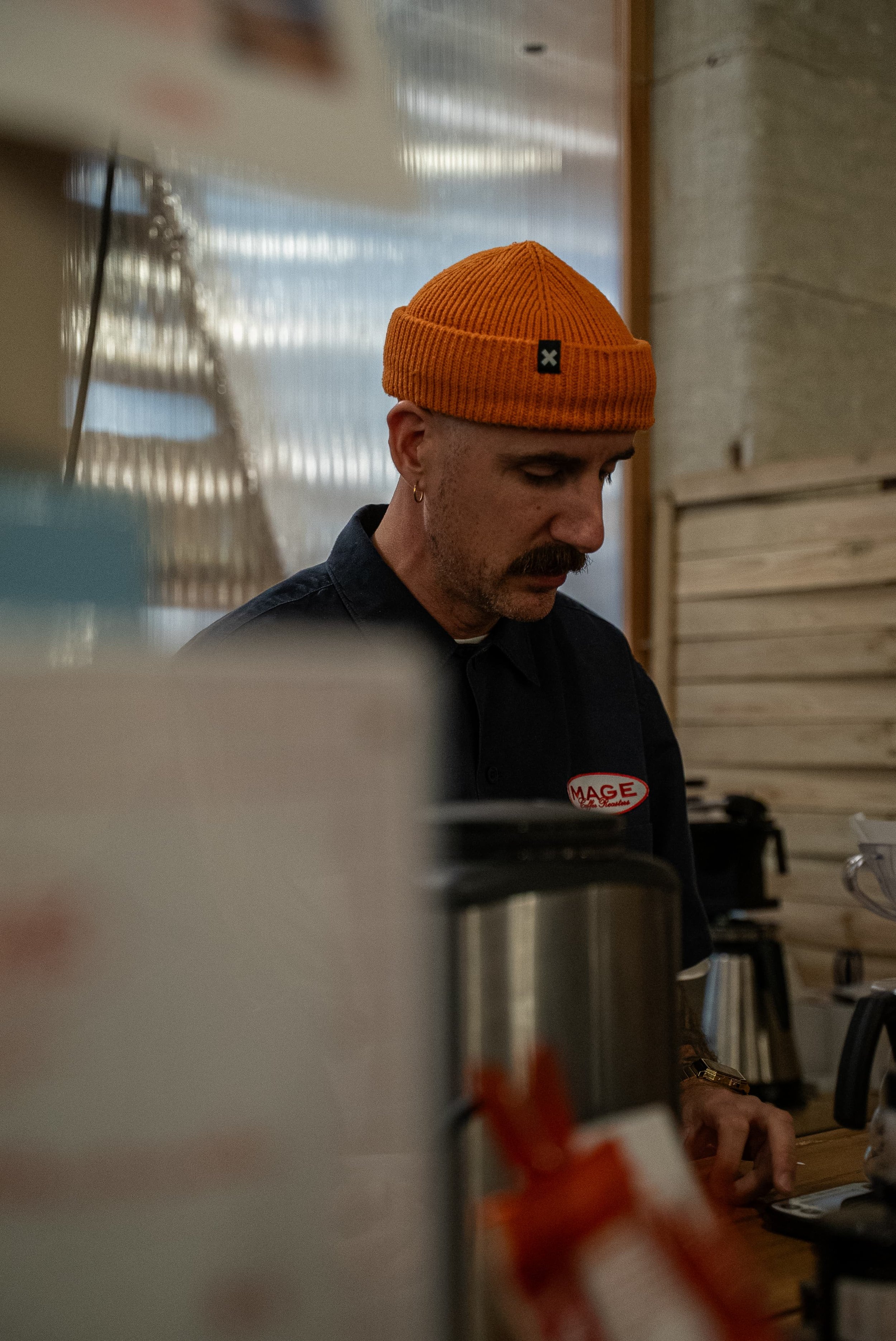 Un homme portant un bonnet orange, vu de profil, travaille dans un environnement intérieur avec des murs en bois et des équipements de cuisine ou de laboratoire autour de lui.
