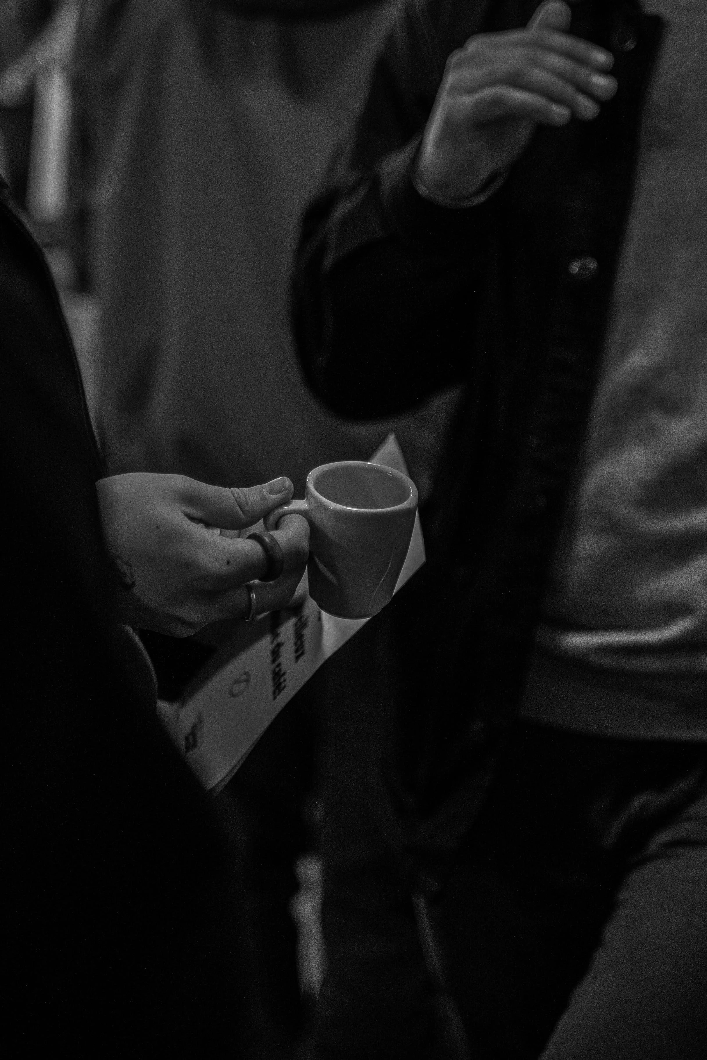 Une personne tient une tasse blanche, l'autre personne tient un smartphone, toutes deux sont partiellement visibles dans une scène sombre.
