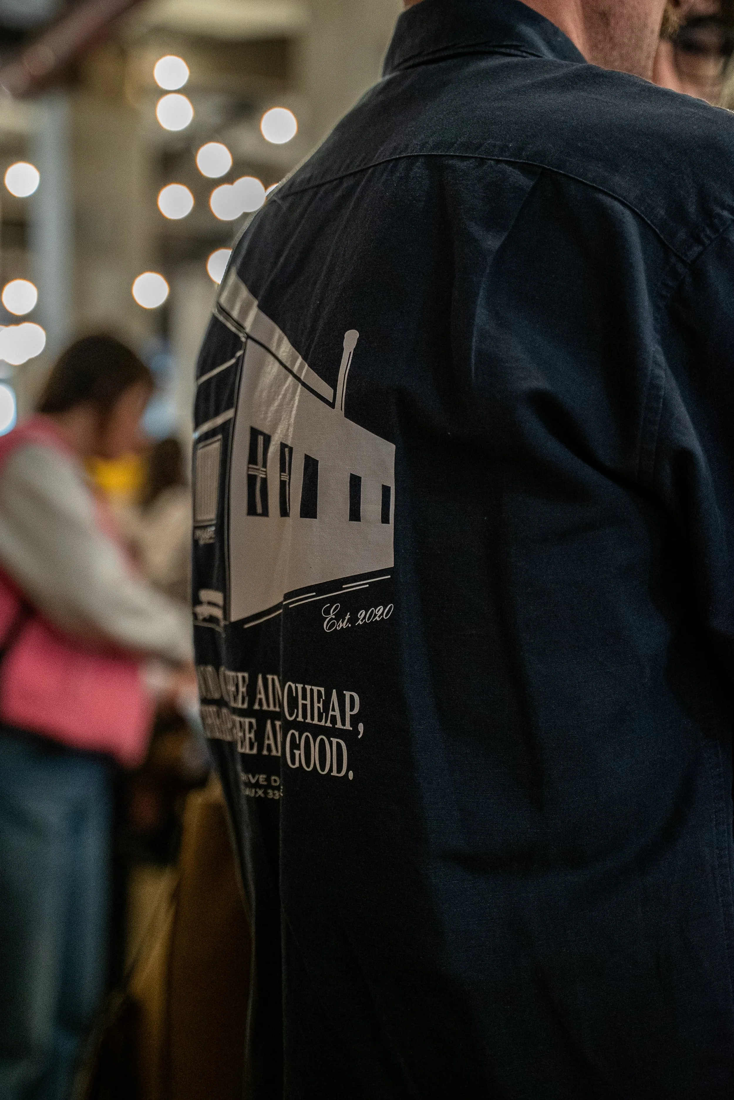 Un homme porte un sweatshirt noir avec un logo de bâtiment blanc et un texte en anglais partiel sur le dos, dans un décor intérieur avec des lumières floues en arrière-plan.