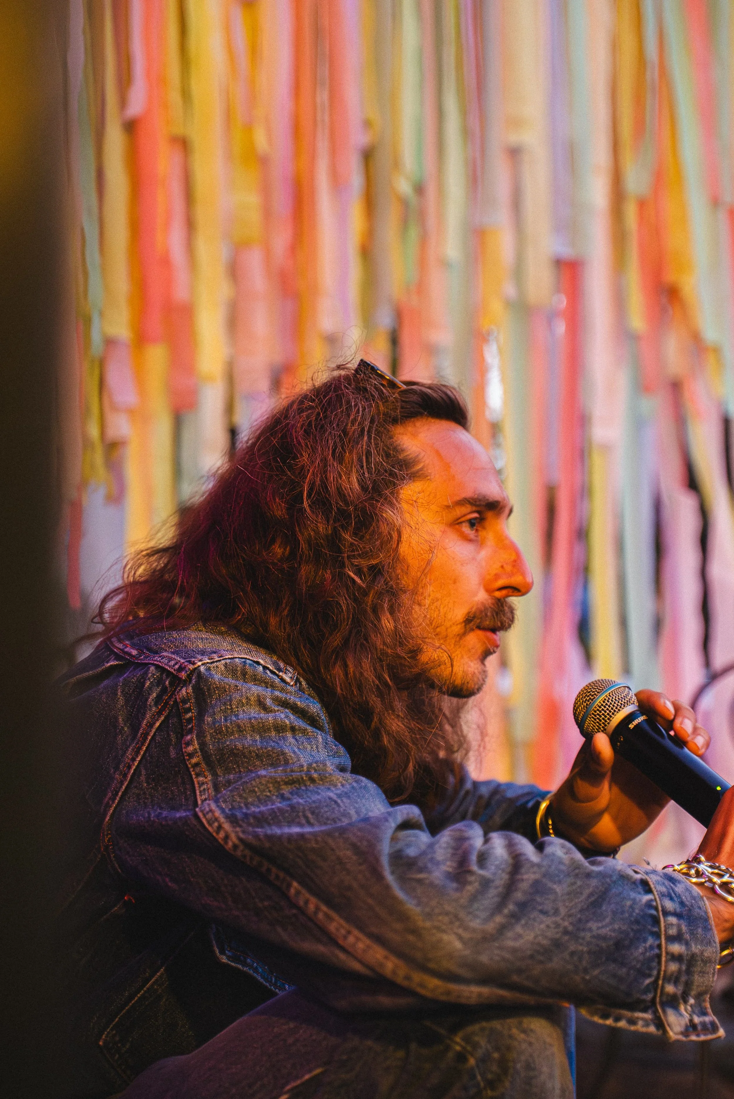 Homme avec longs cheveux et barbe tenant un microphone, portant une veste en jean, au fond un mur coloré avec bandes de tissu.