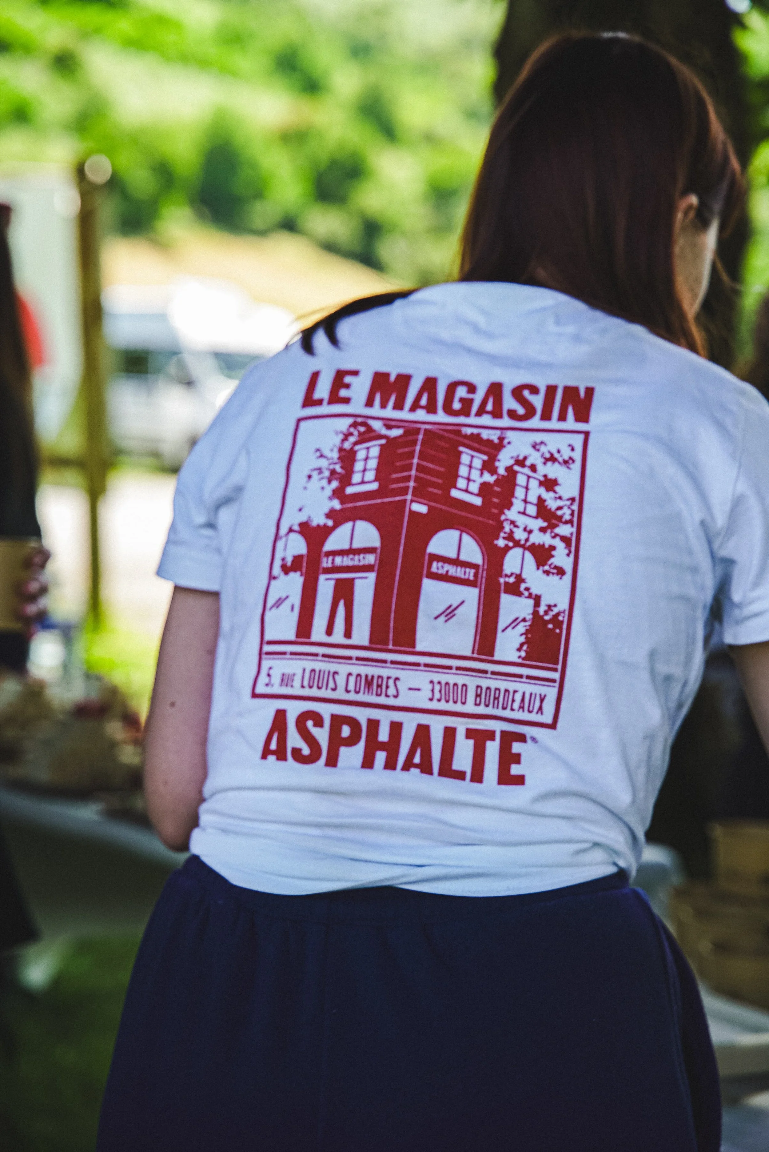 Une personne portant un t-shirt blanc avec des inscriptions en rouge, debout à l'extérieur, dans un environnement verdoyant.