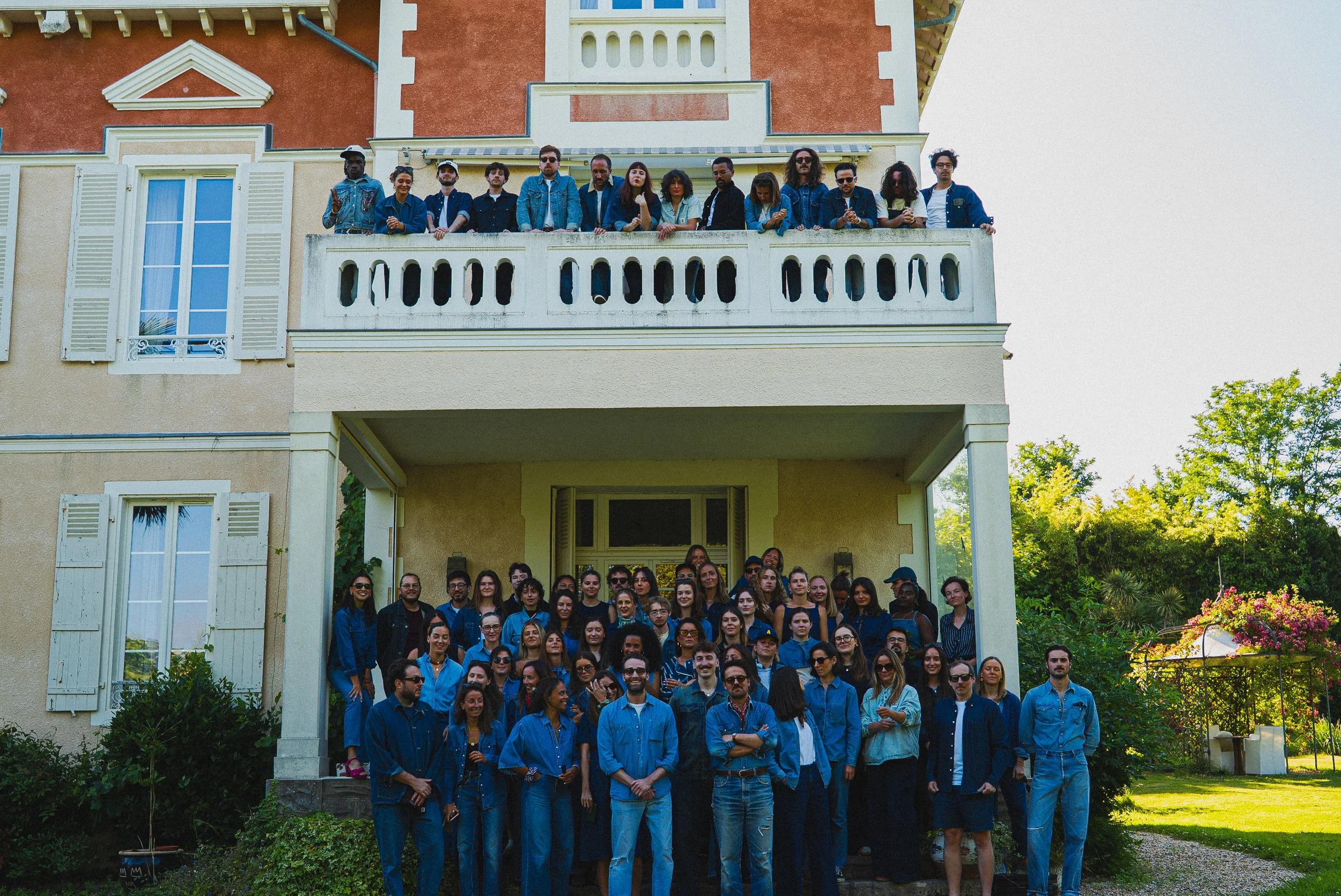 Groupe de personnes posant devant une maison avec un balcon, toutes portent des vêtements bleus, sous un ciel clair et entourées de végétation.