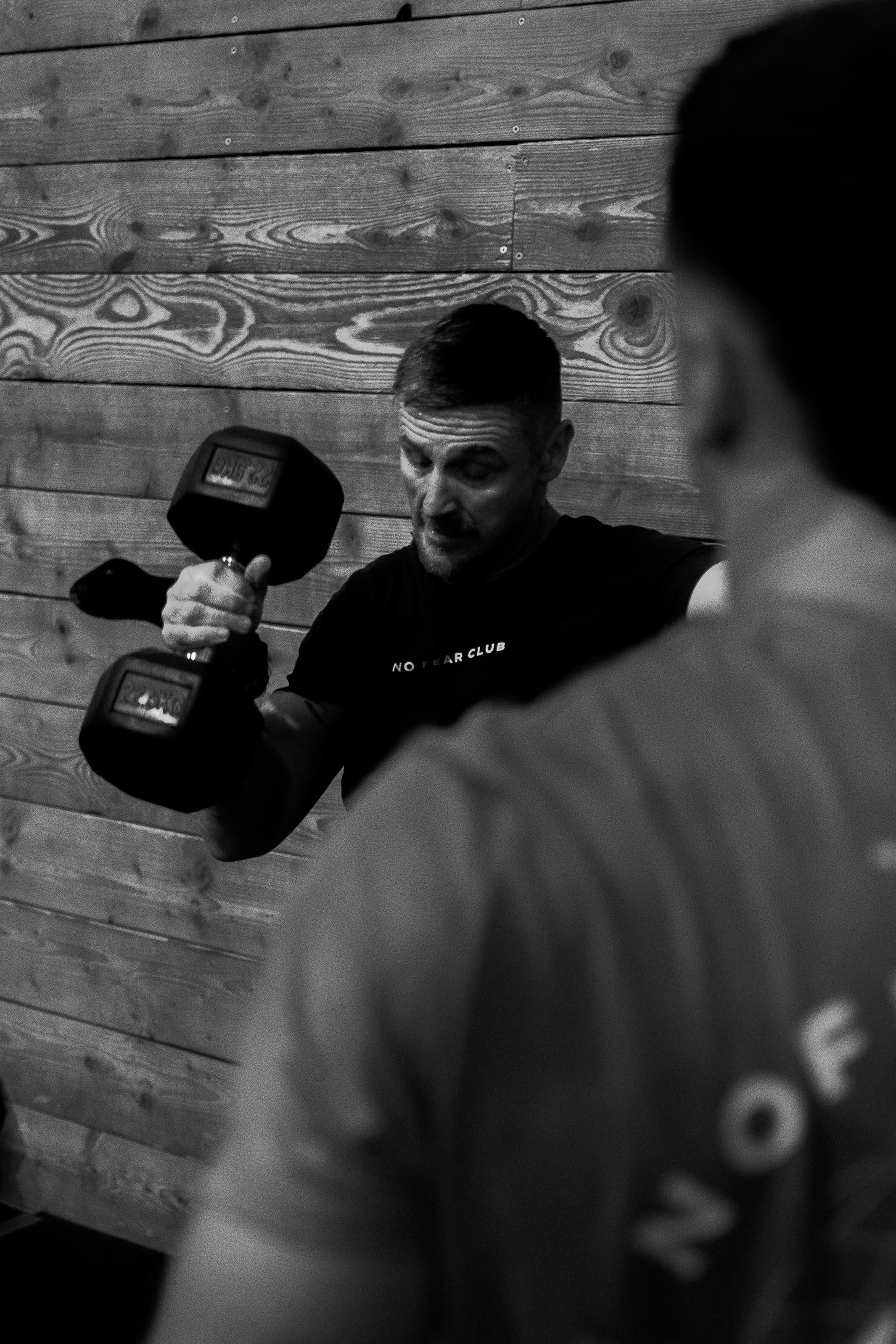 Photo en noir et blanc d'une personne faisant de la musculation avec un haltère, près d'un mur en bois. Perspective partielle floue d'une autre personne dans l'image.