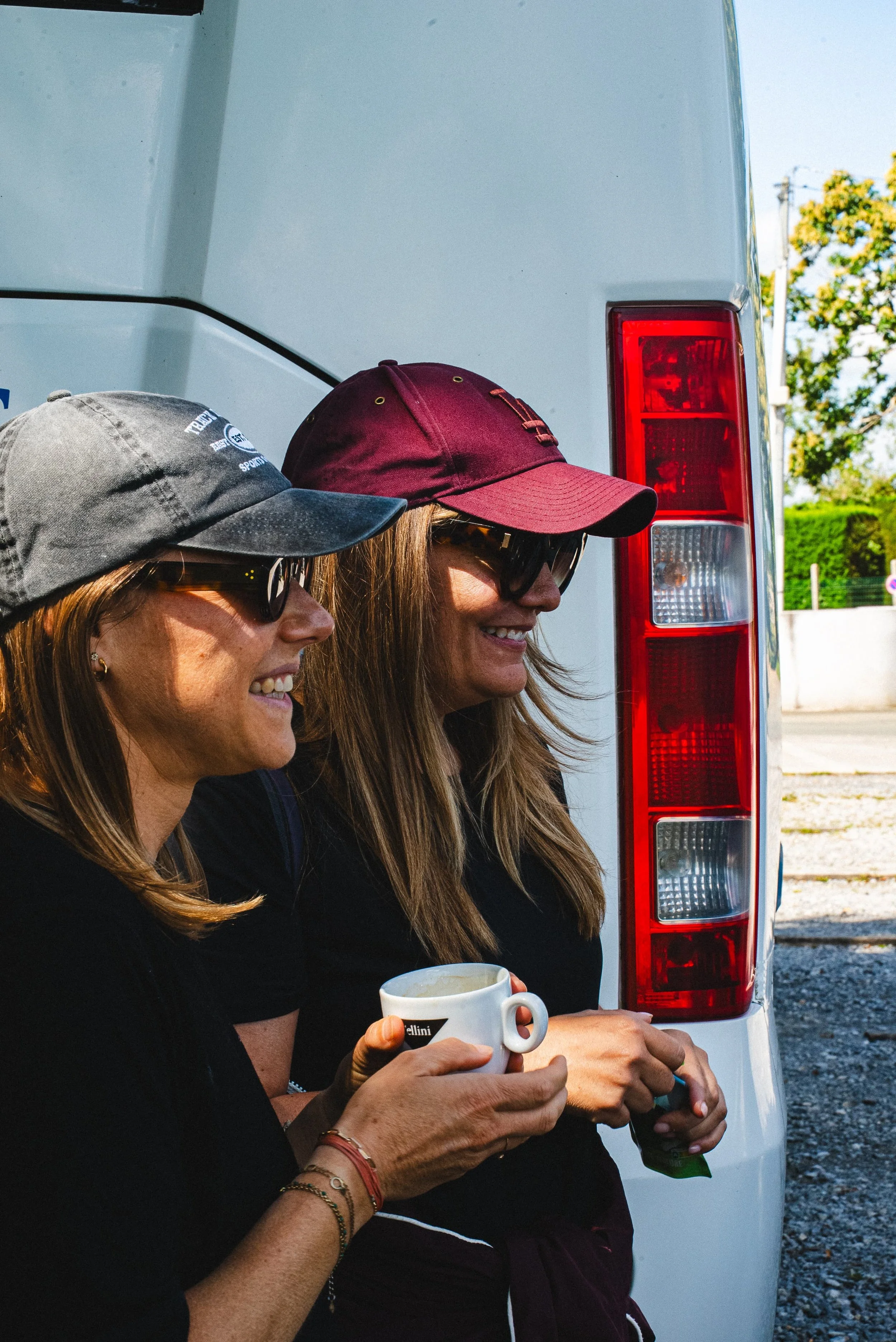 Deux femmes portant des casquettes et des lunettes de soleil, tenant des tasses, debout à côté d'un véhicule blanc, en plein air avec des arbres en arrière-plan.