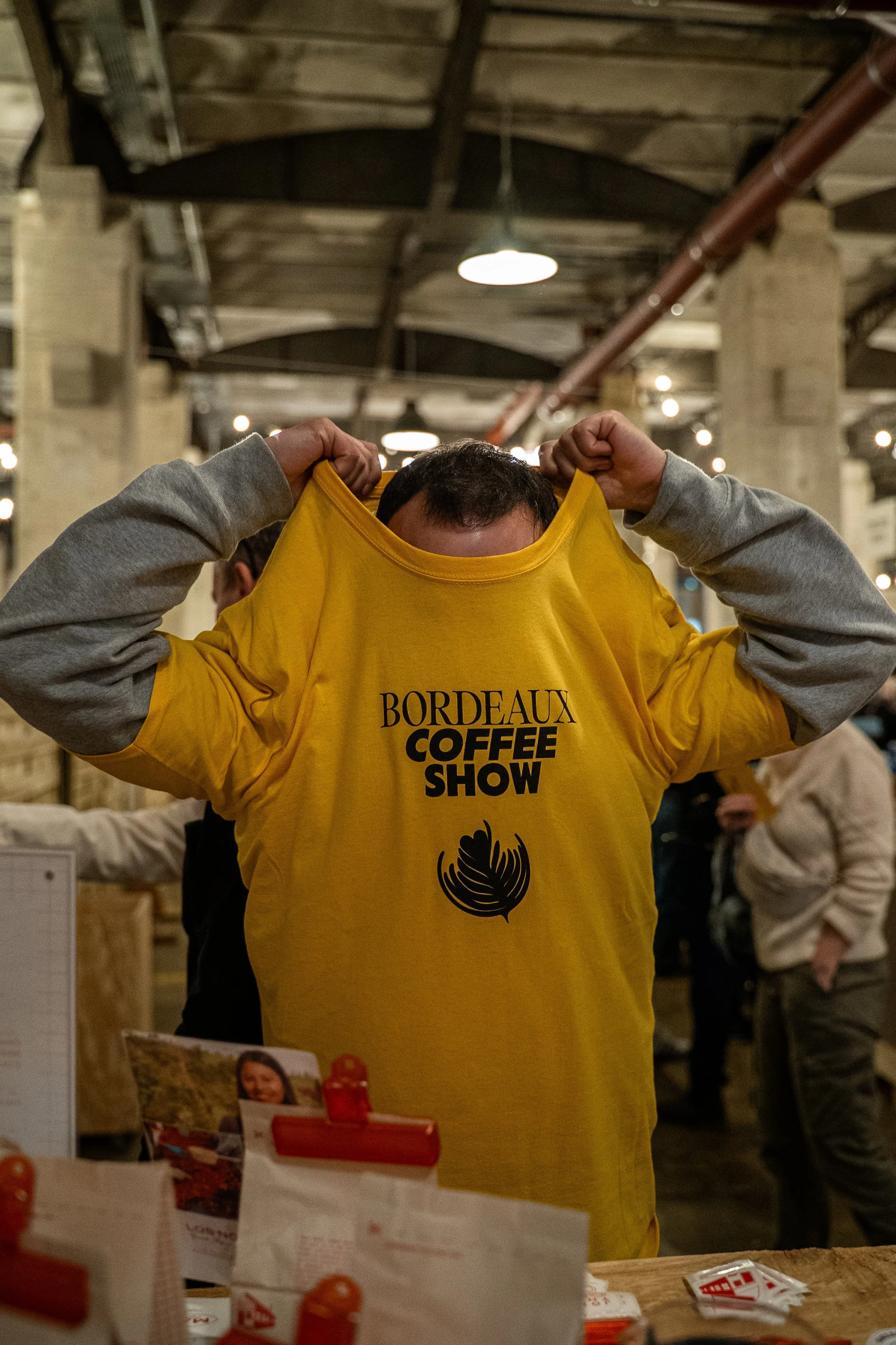 Une personne porte un t-shirt jaune à l'arrière d'un marché couvert, en levant le t-shirt pour révéler son visage. Le t-shirt a l'inscription 'Bordeaux Coffee Show' et une illustration d'une cabosse de cacao.