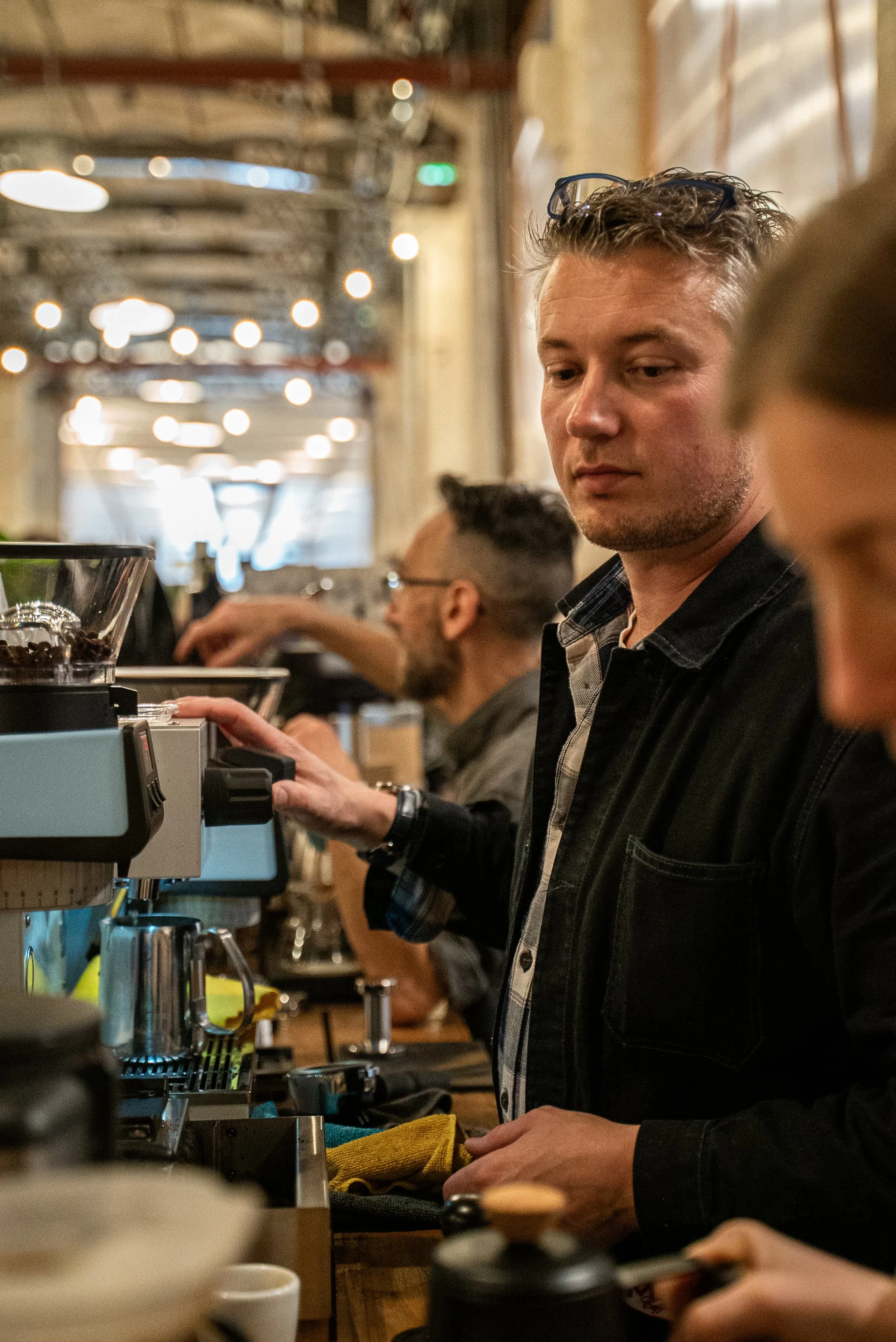 Un homme en train de préparer un café dans un café, avec d'autres personnes en arrière-plan, ambiance chaleureuse et éclairage tamisé.