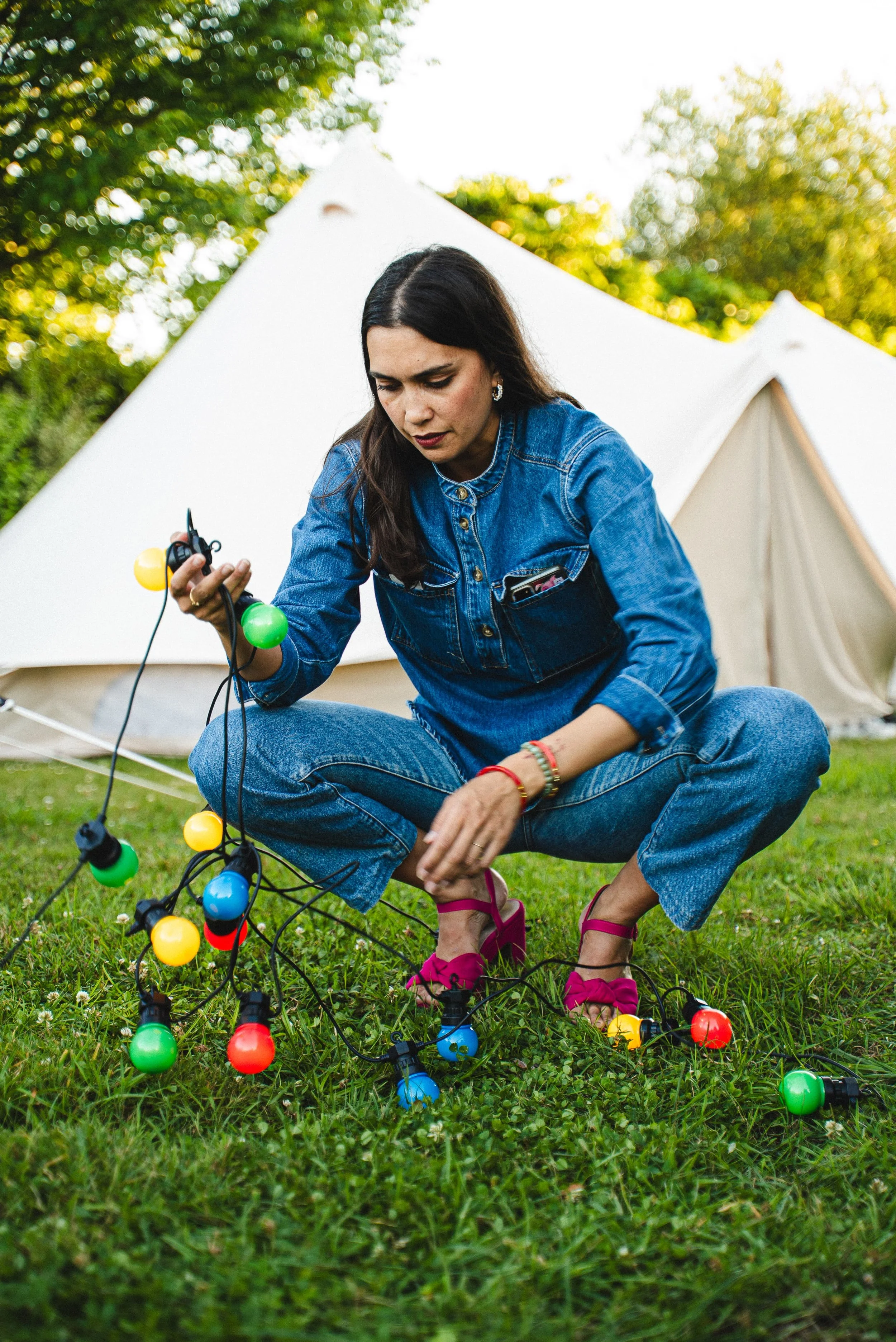 Femme assise dans l'herbe, manipulant des guirlandes lumineuses colorées, avec une tente blanche en arrière-plan, sous un ciel ensoleillé.
