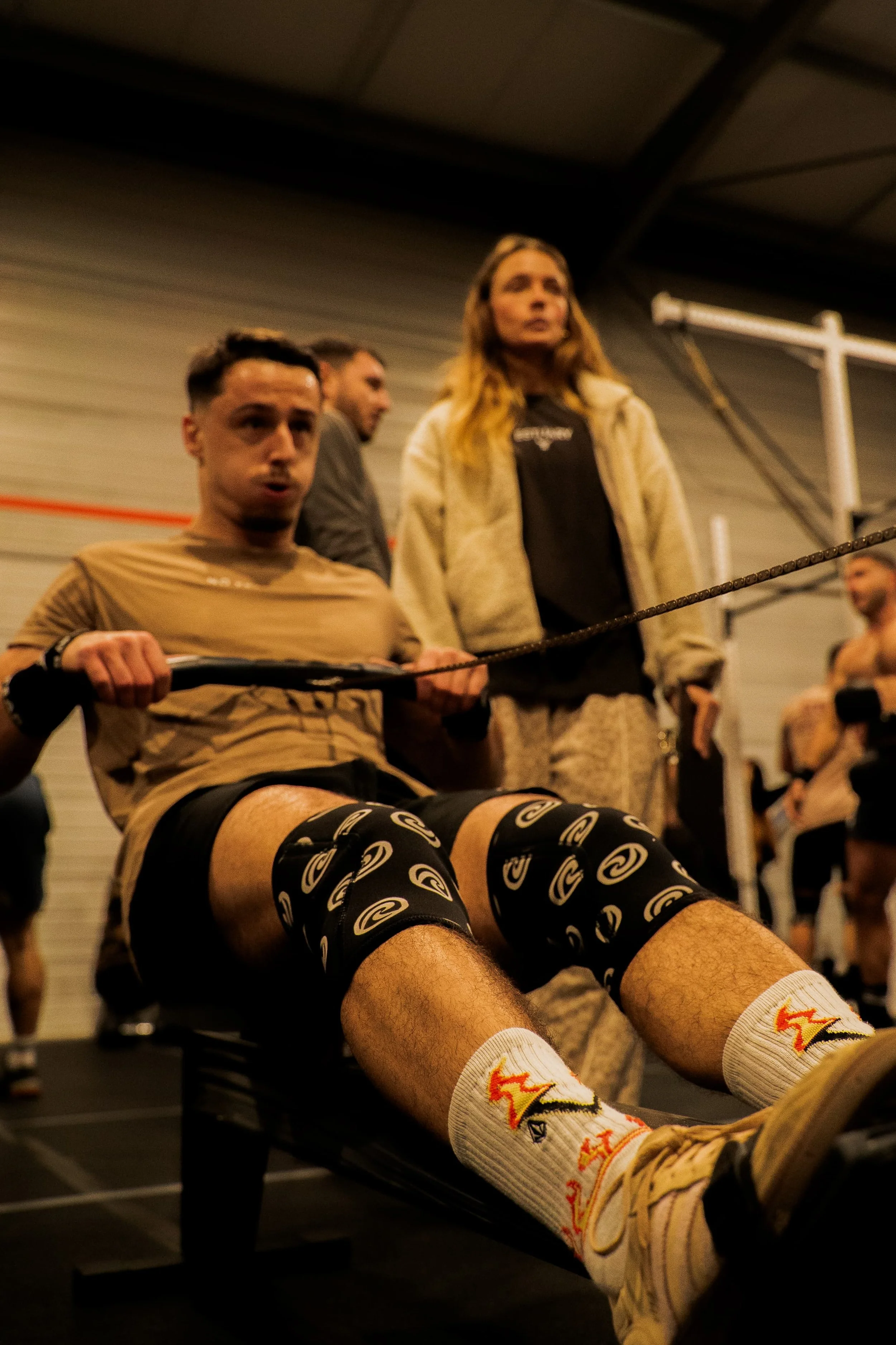 Un homme assis sur un banc de musculation, portant des chaussettes avec un logo. Il est dans une salle de sport, entouré d'autres personnes, avec des murs en métal et un plafond industriel.