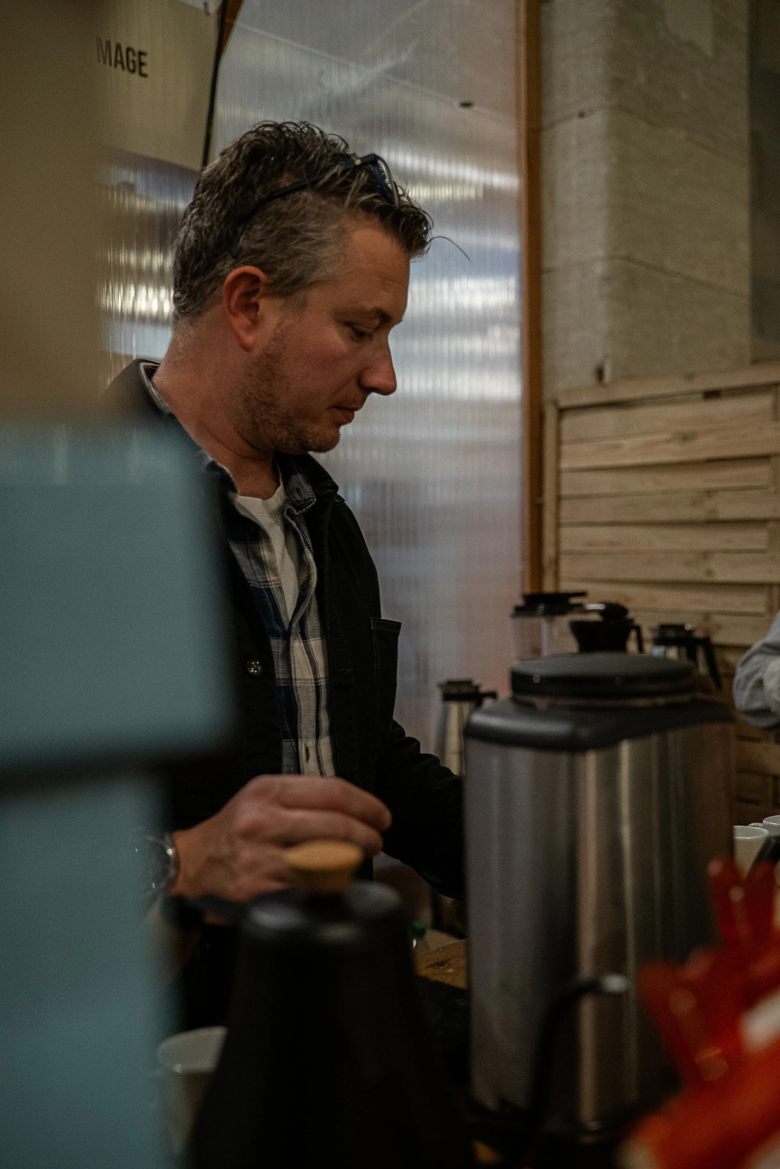 Un homme avec cheveux bruns courts et barbe légère, portant une chemise à carreaux et un blouson, se tient à côté d'une machine à café dans un espace intérieur en bois, regardant vers le bas.