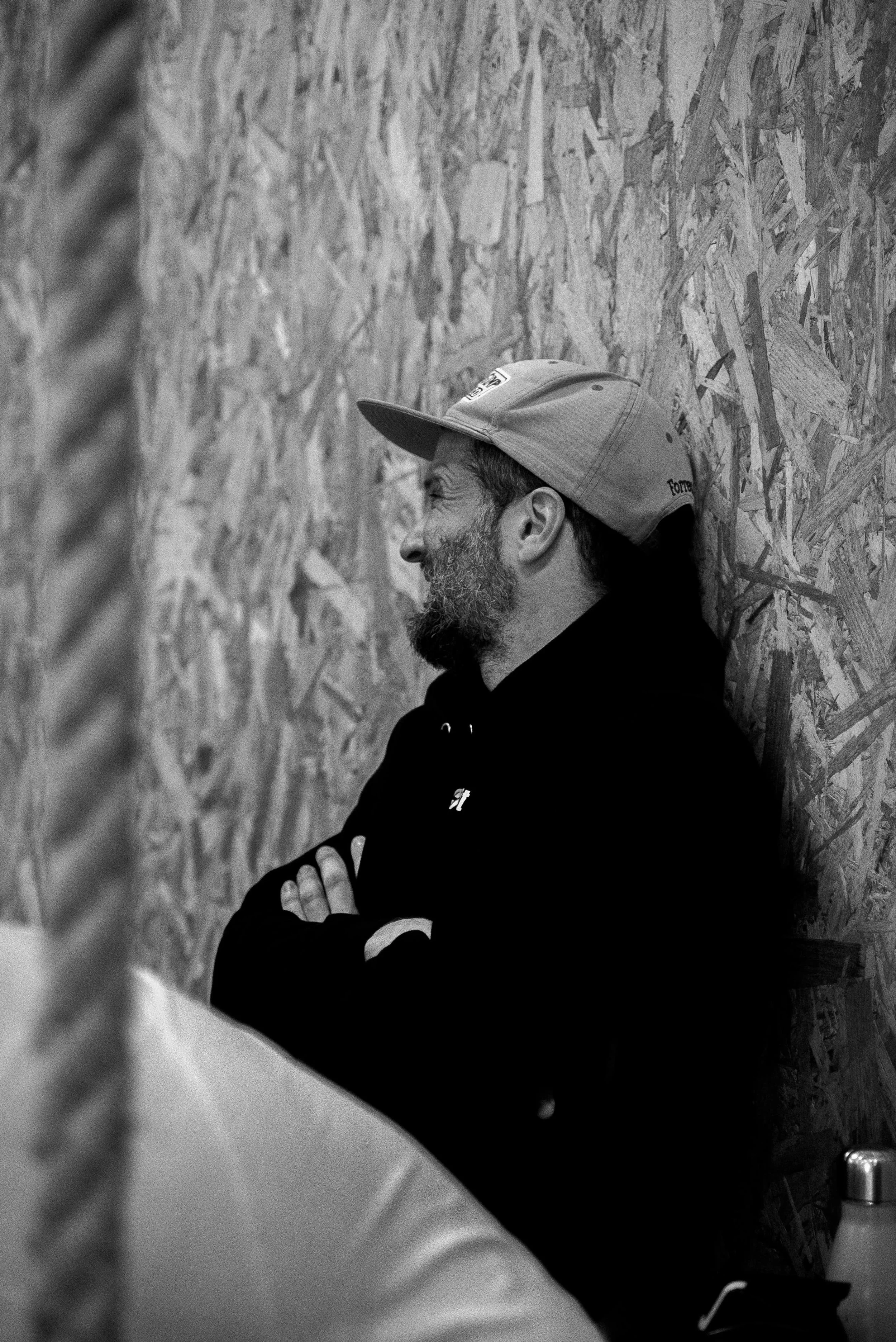 Un homme barbu portant une casquette et un sweat noir, debout contre un mur en panneaux de bois mélangés, souriant.