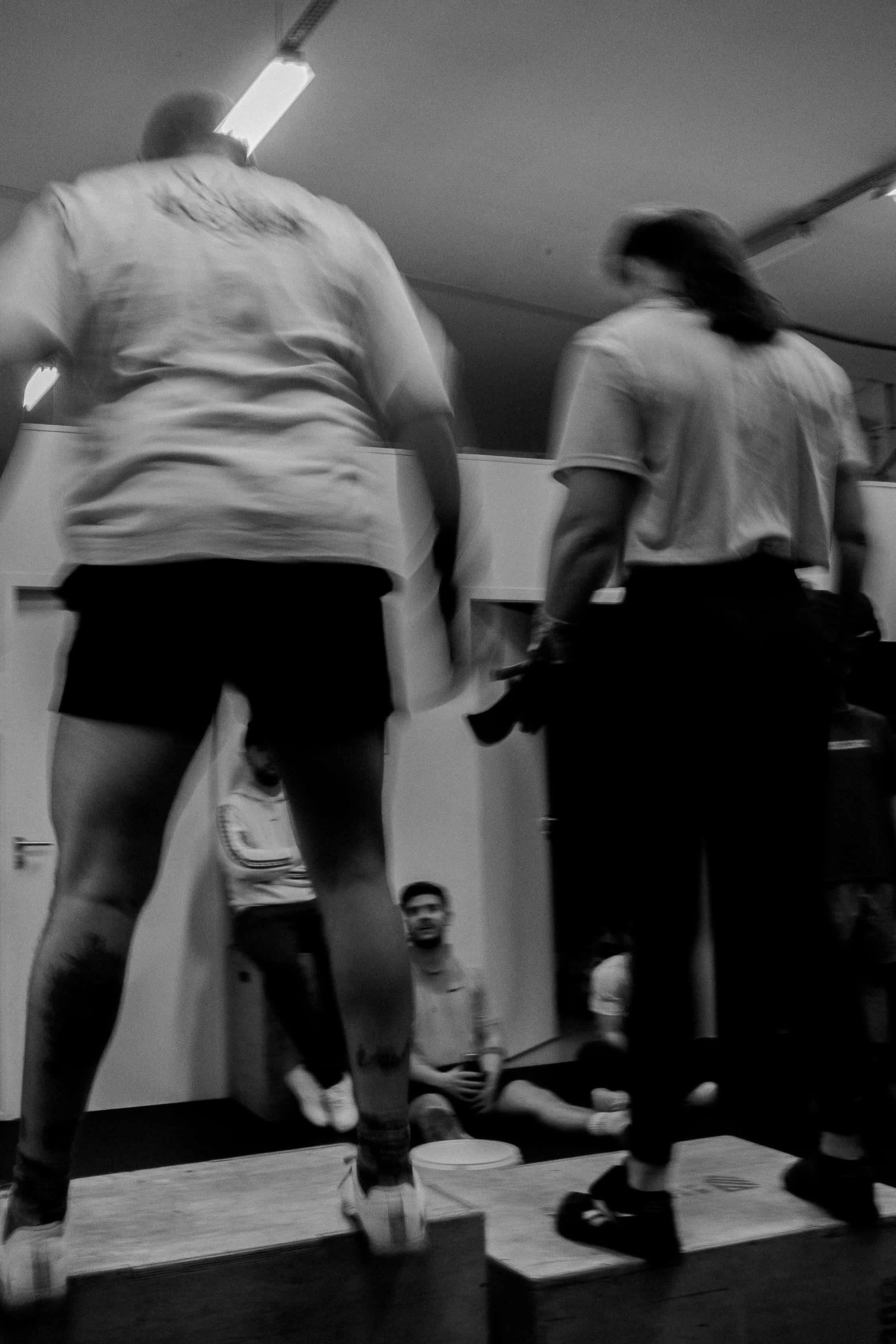 Deux personnes en tenue de sport effectuent un saut sur une plateforme en bois, dans un environnement intérieur. Plusieurs autres personnes en arrière-plan regardent l'activité.