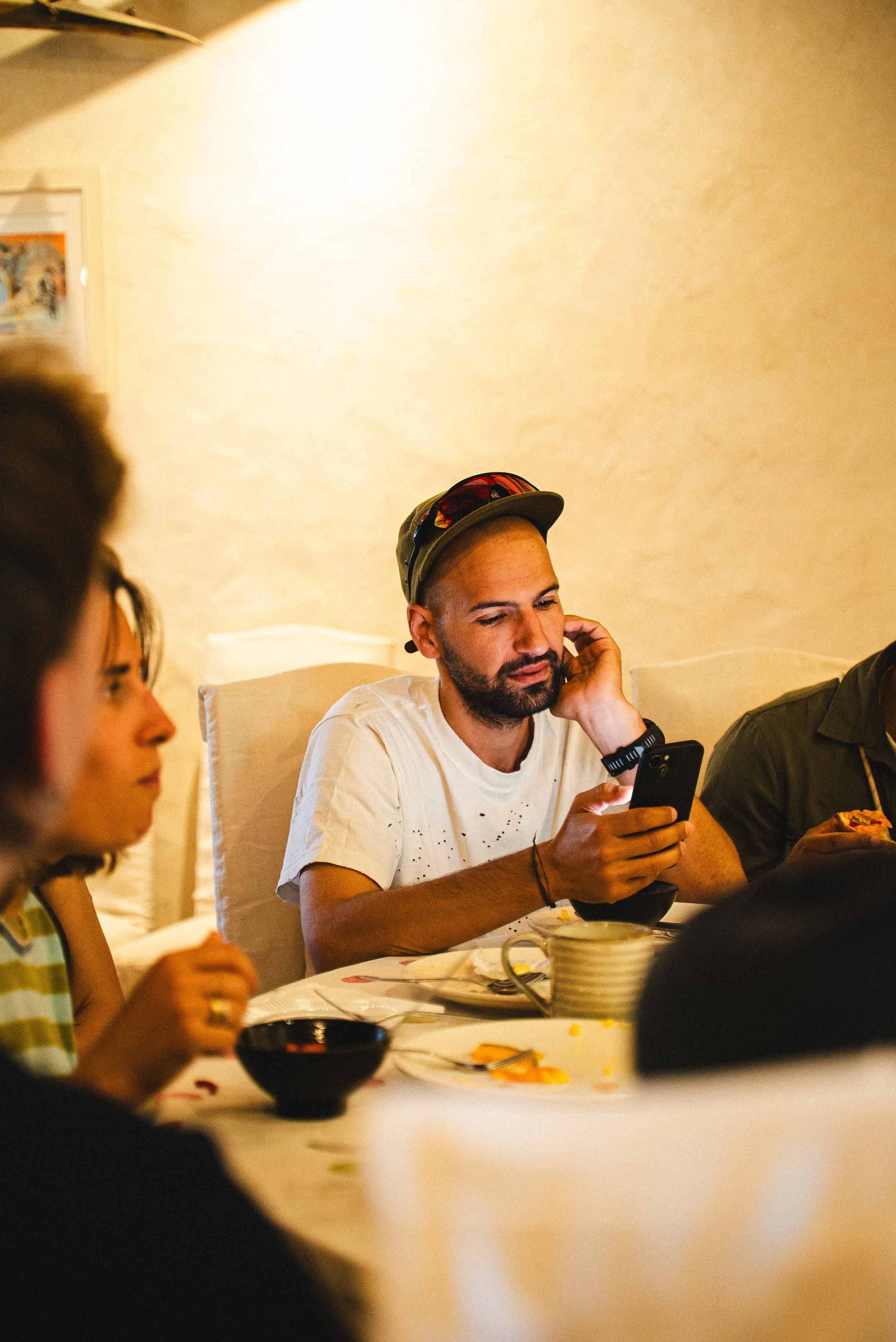 Un groupe de personnes assises autour d'une table lors d'un repas ou d'une réunion, avec un homme portant une casquette et regardant un téléphone portable en premier plan.