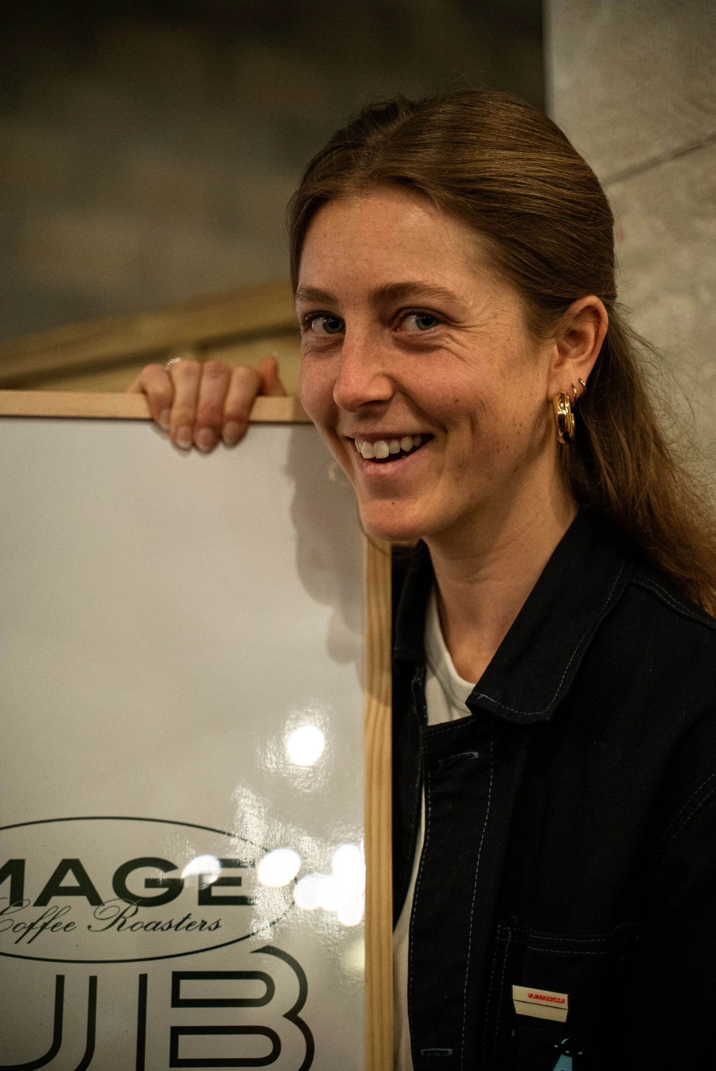 Une femme souriante avec des boucles d'oreilles en or, tenant une planche en bois près d'un panneau de signalisation noir et blanc, dans un intérieur éclairé.