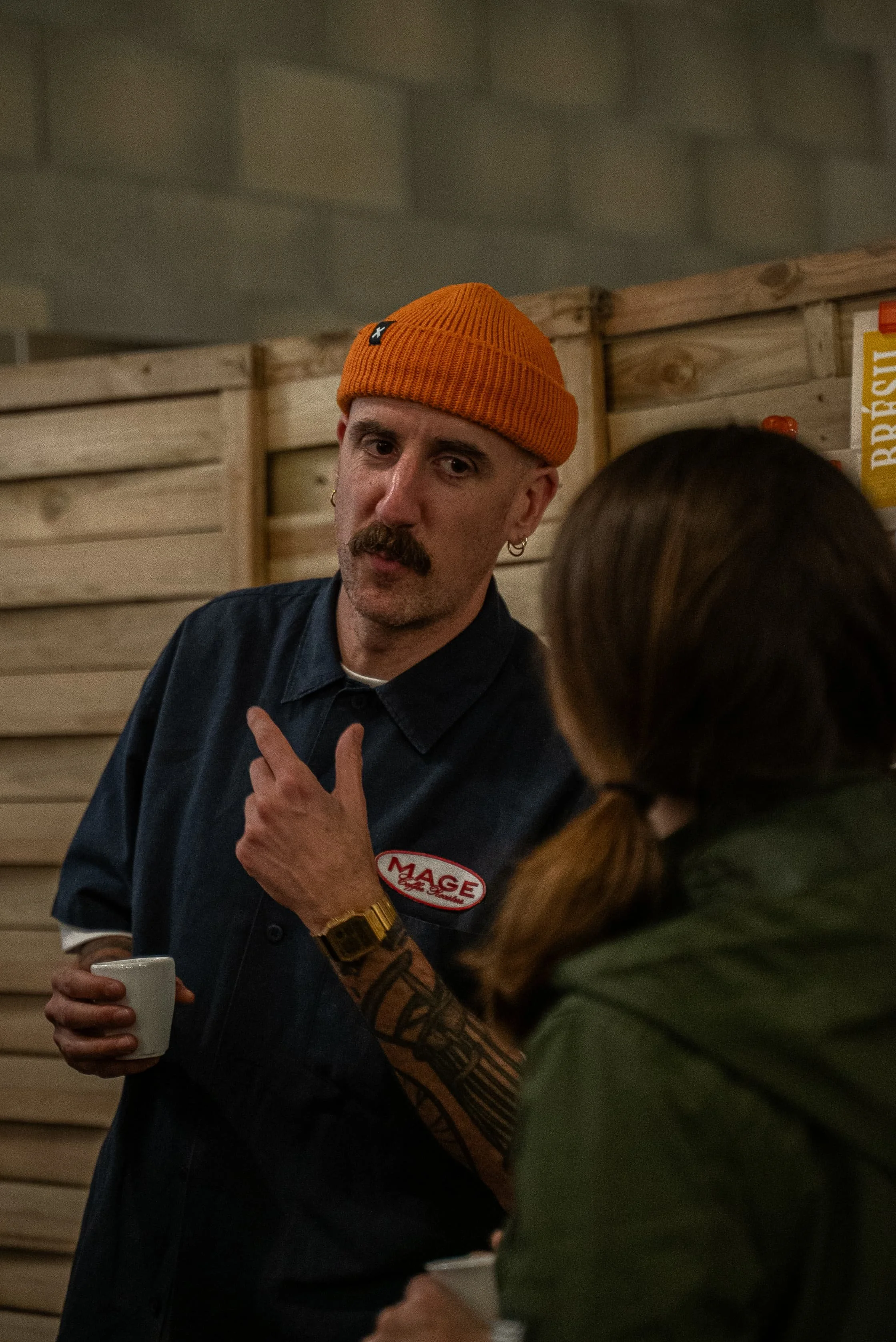 Un homme avec une moustache, portant un bonnet orange et une veste sombre, tient une tasse blanche dans une main et semble discuter avec une femme dont la tête est partiellement visible, dans un environnement intérieur en bois.