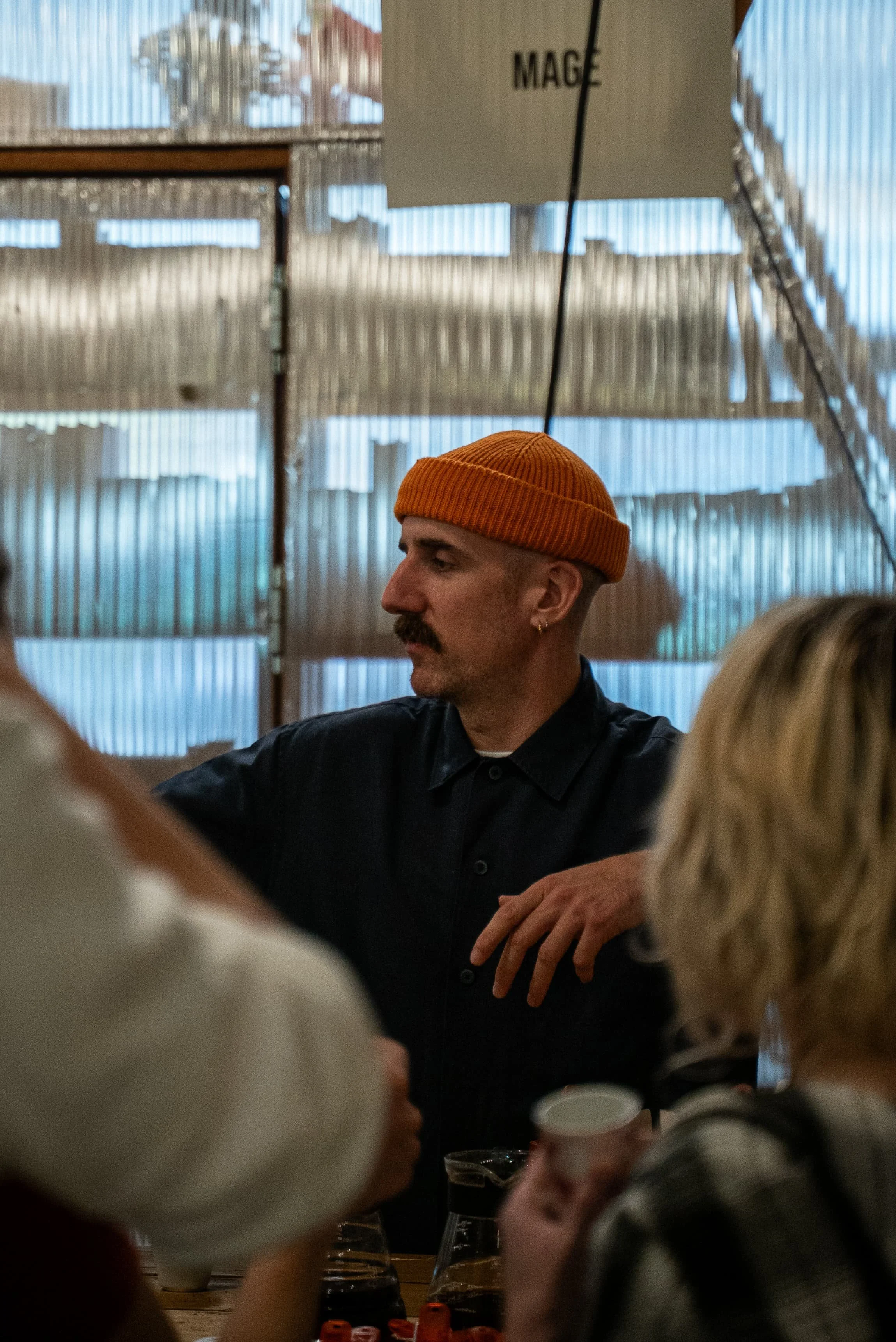 Un homme portant un bonnet orange assis à une table dans un restaurant ou un café, entouré de plusieurs personnes. La scène se déroule à l'intérieur avec un décor en bois et des panneaux en fibre de verre transparents.