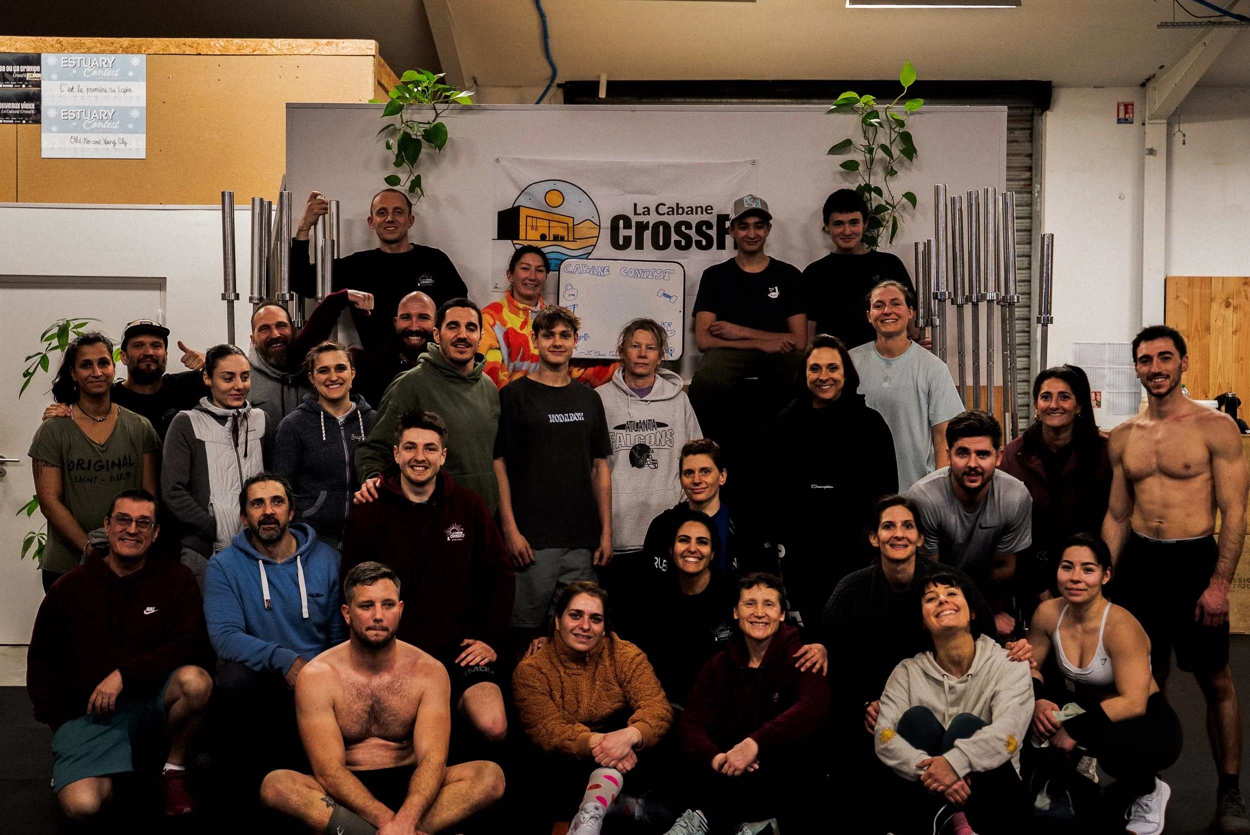 Groupe de personnes posant pour une photo dans une salle, avec un panneau indiquant 'La Cabane CrossFit' en arrière-plan. Certaines personnes sont en tenue de sport, d'autres en vêtements décontractés, et quelques-uns sont assis ou à genoux.