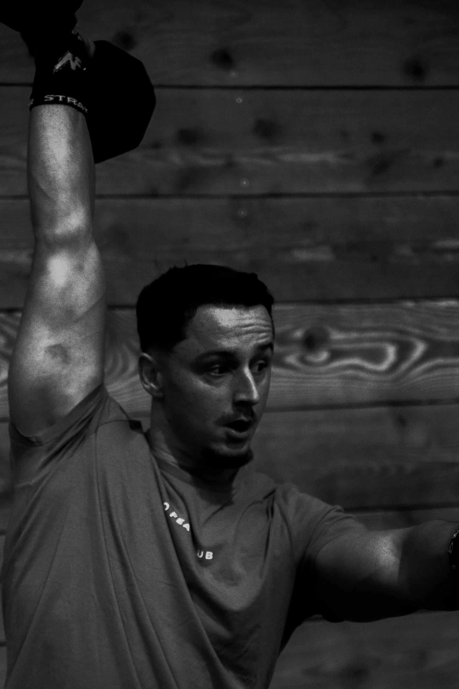 Un homme prépare à faire un exercice ou un mouvement physique, portant un t-shirt et une montre connectée, dans un environnement intérieur avec un plafond en bois.