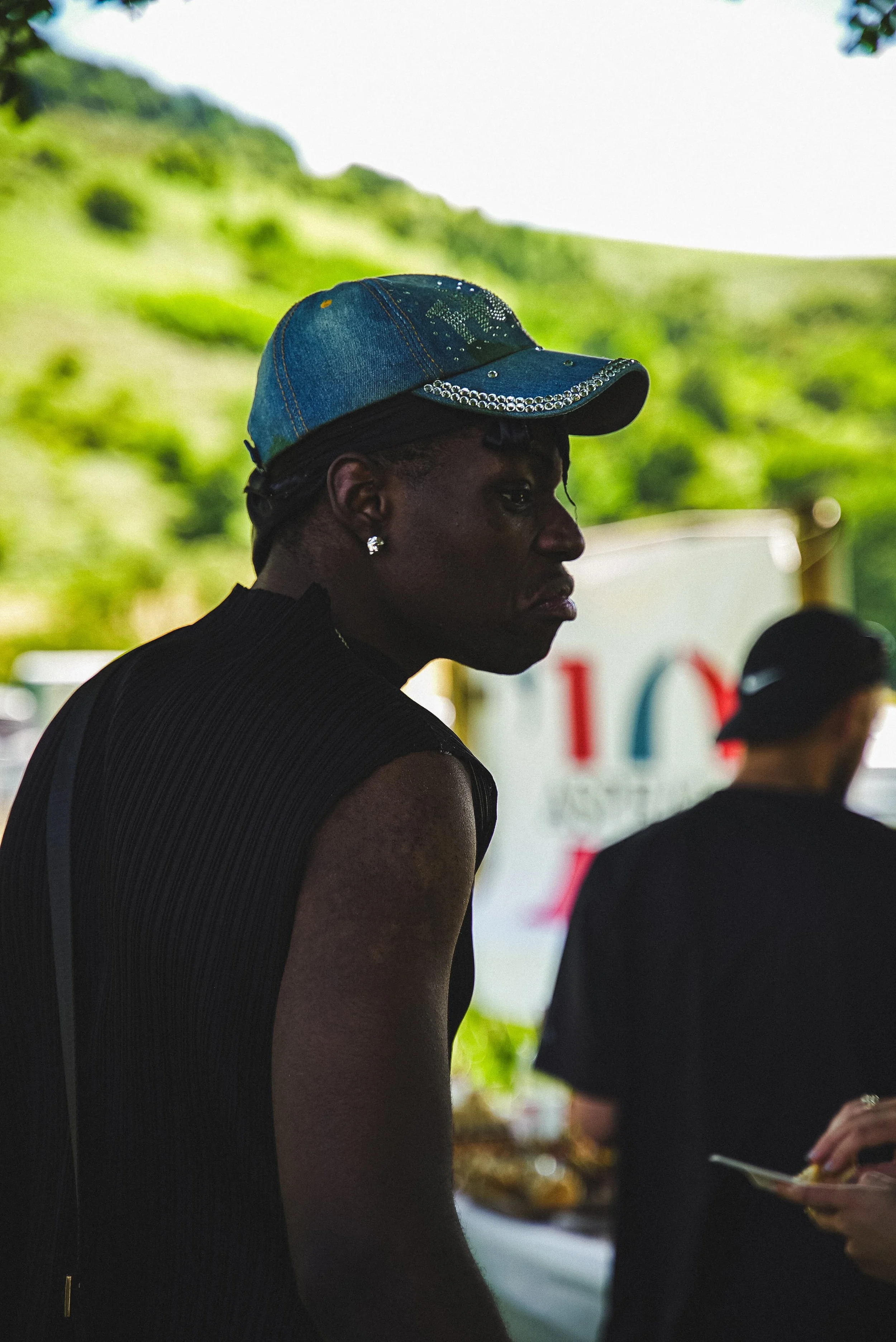 Une femme noire portant une casquette en jean décorée de rhinestones, vue de profil, dans un environnement extérieur avec un fond verdoyant.