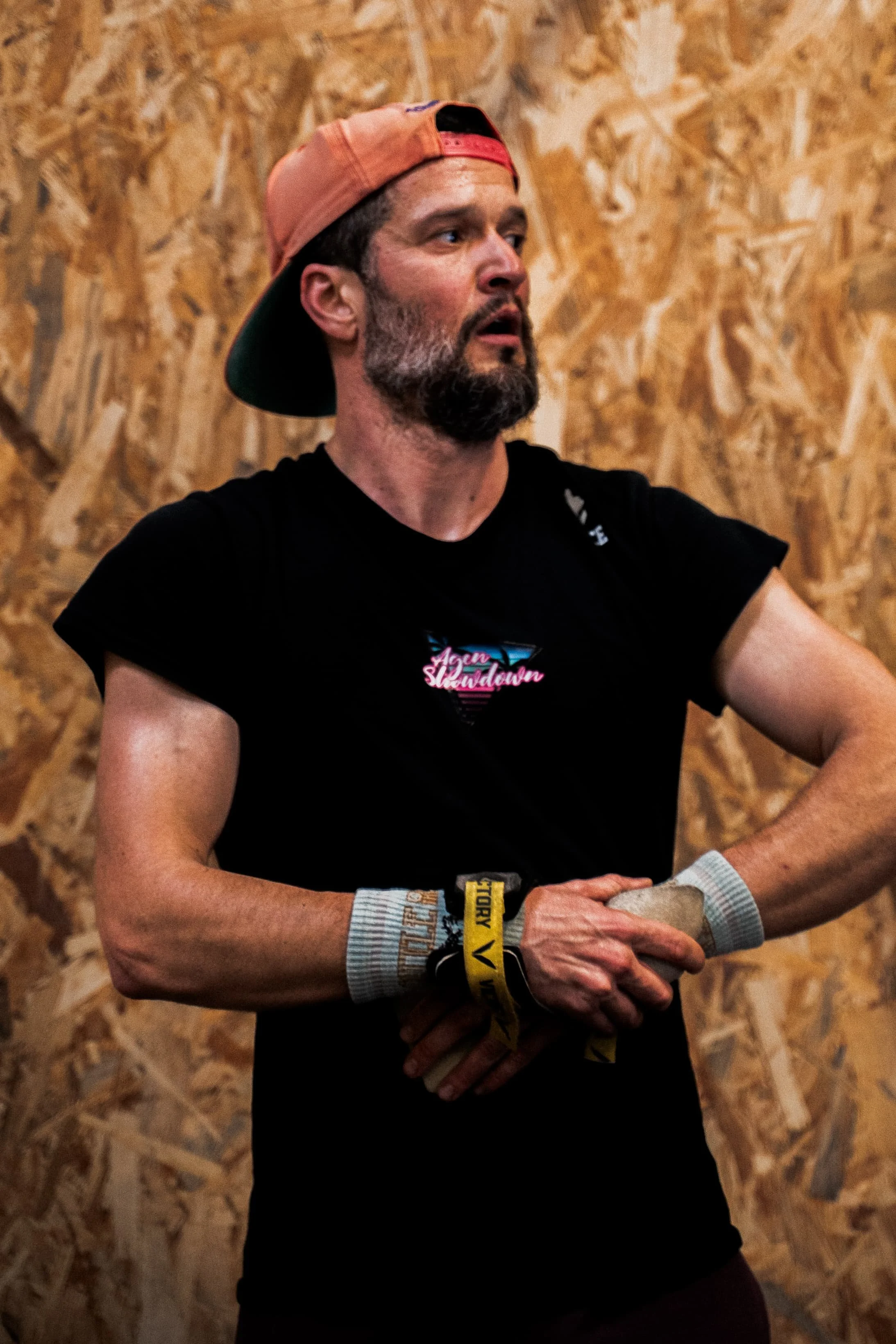 Homme avec barbe, portant une casquette à l'envers et un t-shirt noir, assis à une table en bois contre un mur en panneau de particules, avec un bracelet de course au poignet gauche.