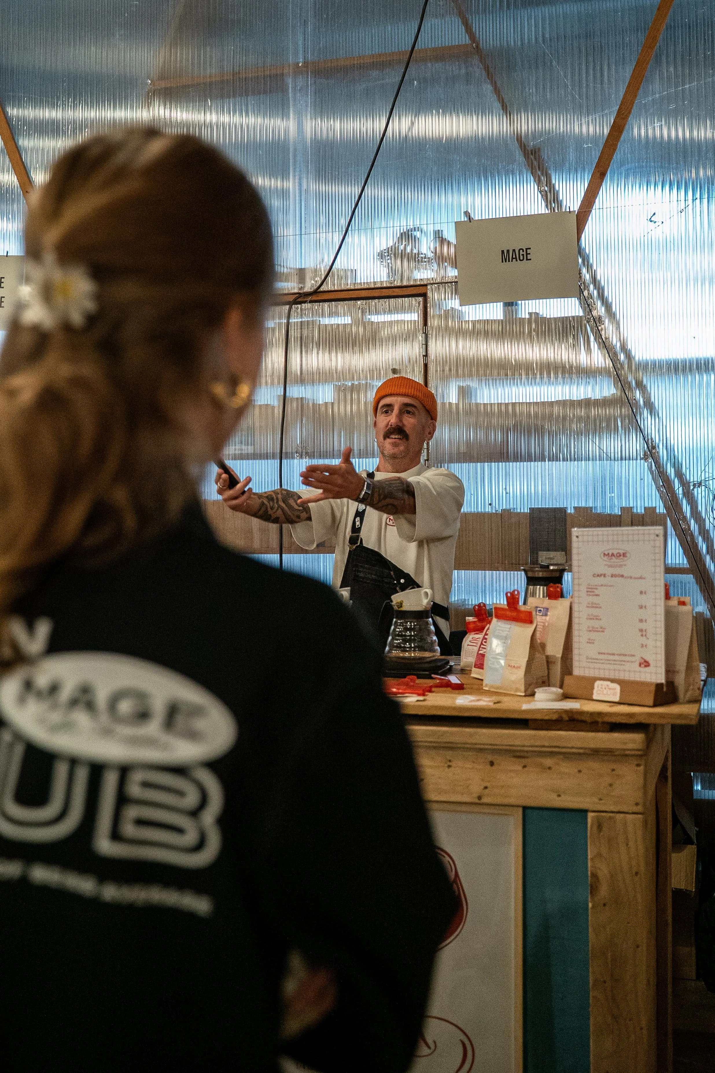 Un homme portant un bonnet orange et un t-shirt blanc, parlant ou expliquant quelque chose à une personne dans un café ou un stand de nourriture, avec une enseigne indiquant 'MAGE' et divers articles de menu sur la table.