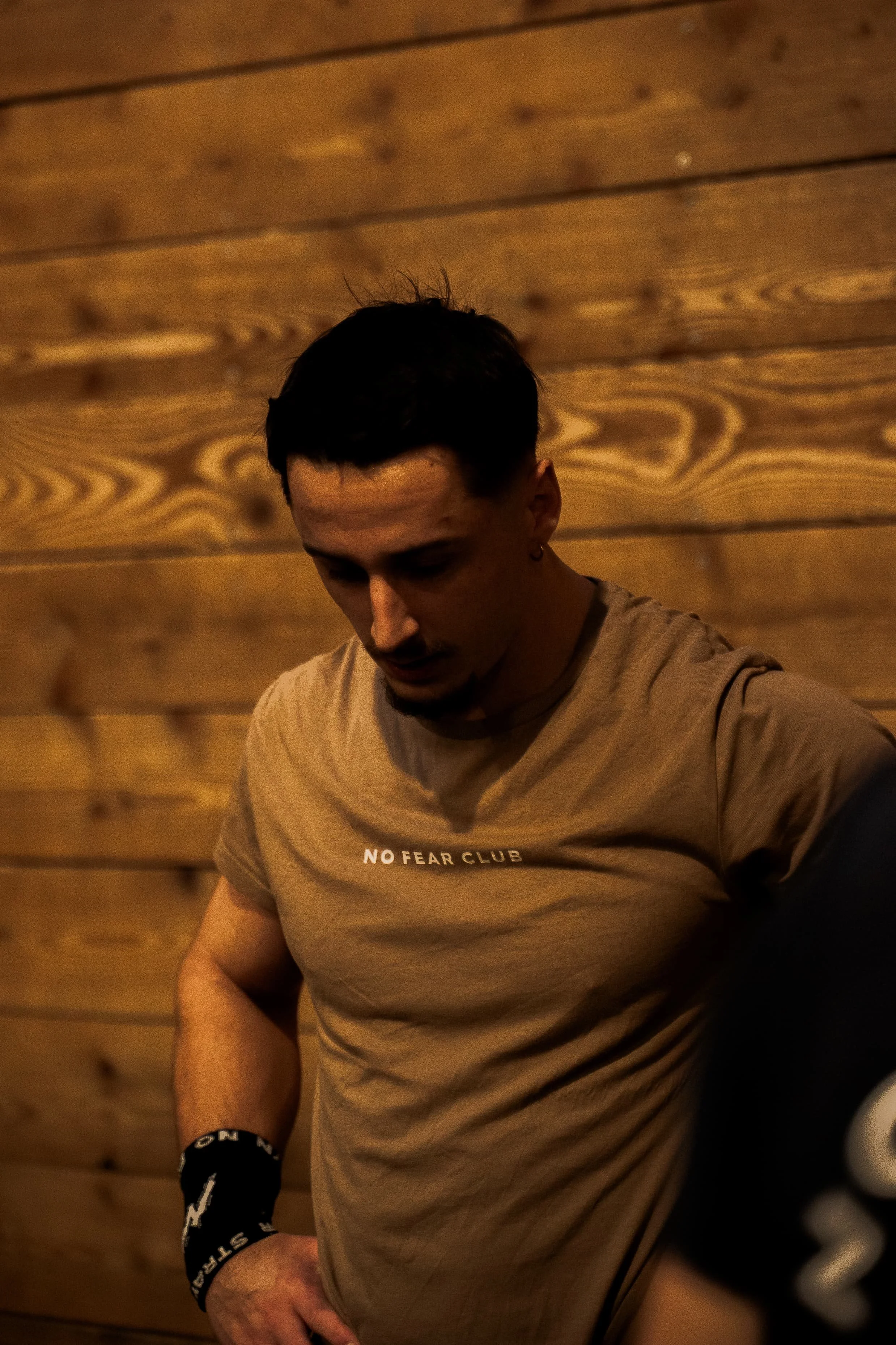 Jeune homme avec cheveux noirs, portant un t-shirt beige avec l'inscription "NO FEAR CLUB" et un bracelet noir, assis contre un mur en bois.