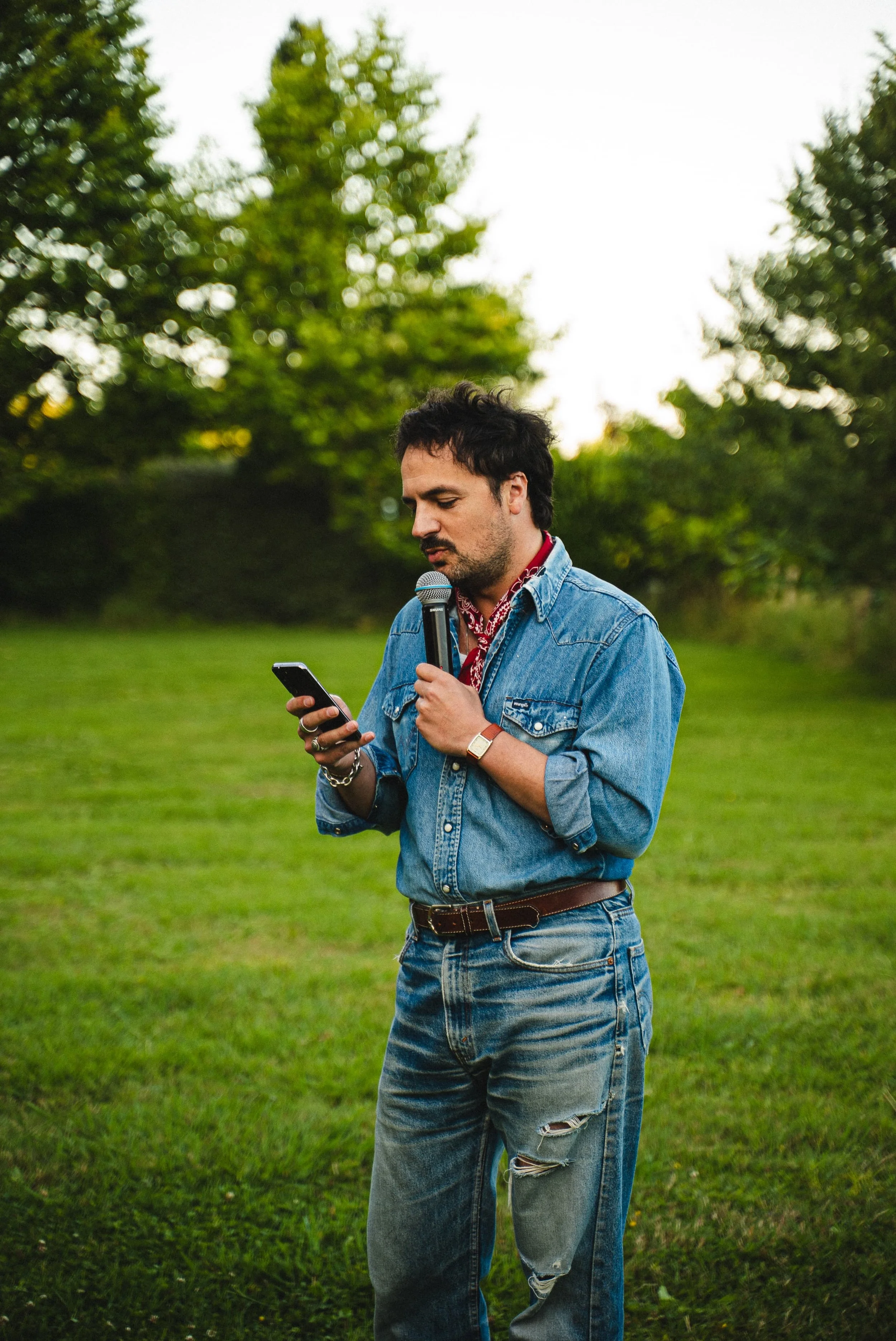 Un jeune homme avec des cheveux bouclés noirs, portant une chemise en denim et un bandana rouge autour du cou, tient un microphone dans une main et un téléphone dans l'autre, se trouvant dans un parc verdoyant avec des arbres en arrière-plan.